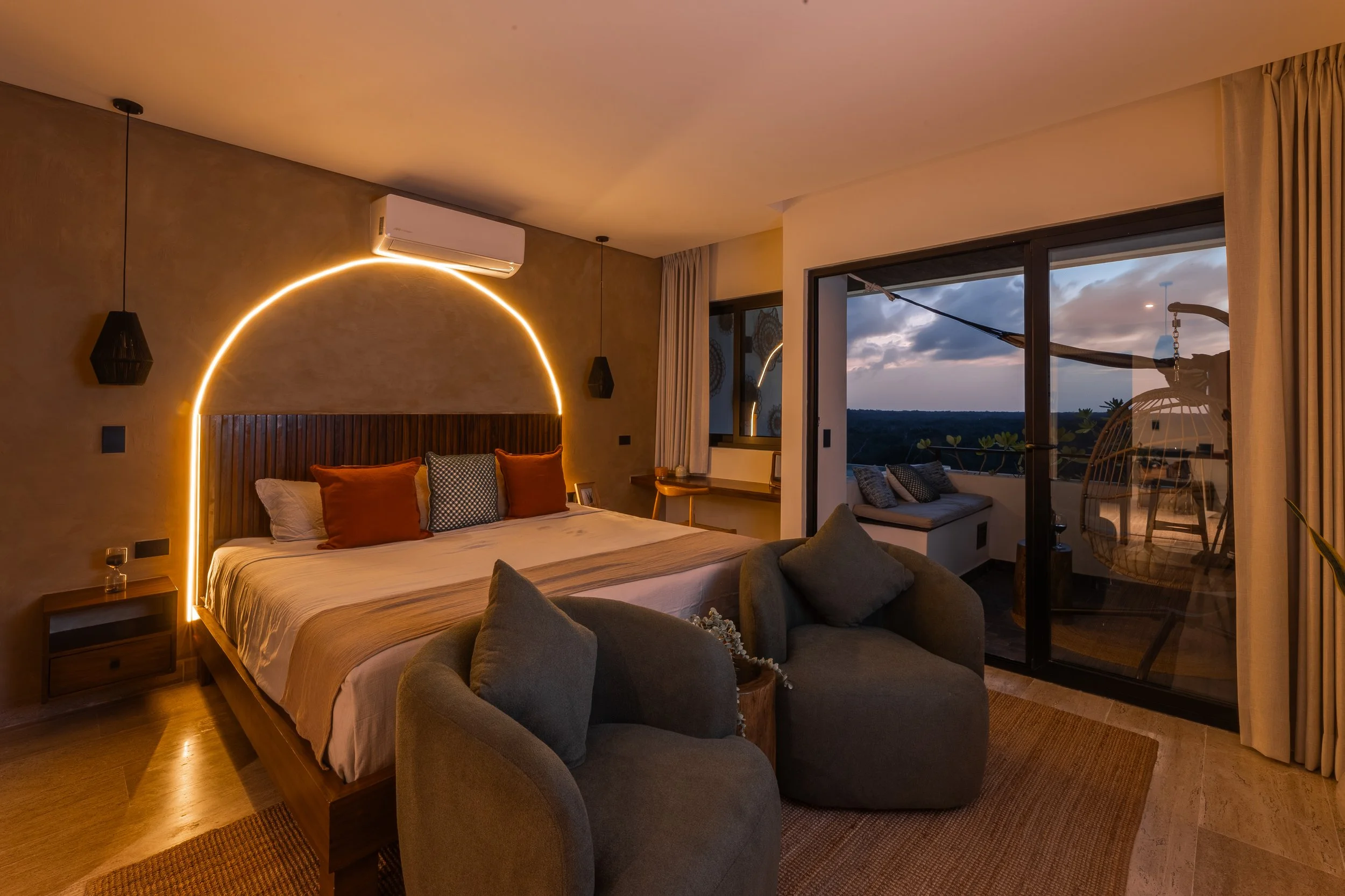 A modern bedroom with a wooden bed frame, orange and patterned pillows, a beige throw blanket, and a curtain-covered window showing an evening sky with clouds, at Mahahual.