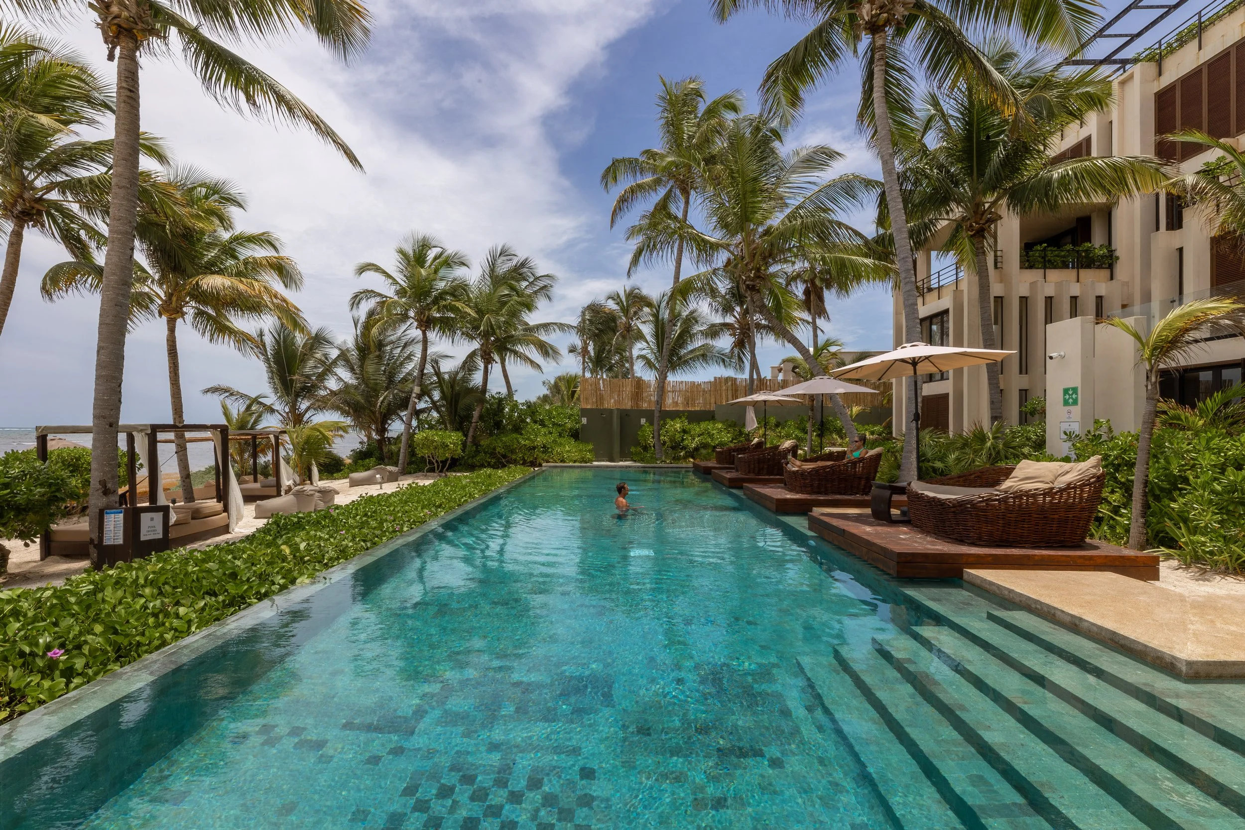 A tropical pool area surrounded by palm trees and a building with balconies. There are lounge chairs with umbrellas, and a person swimming in the pool, at Bespoke Tulum.