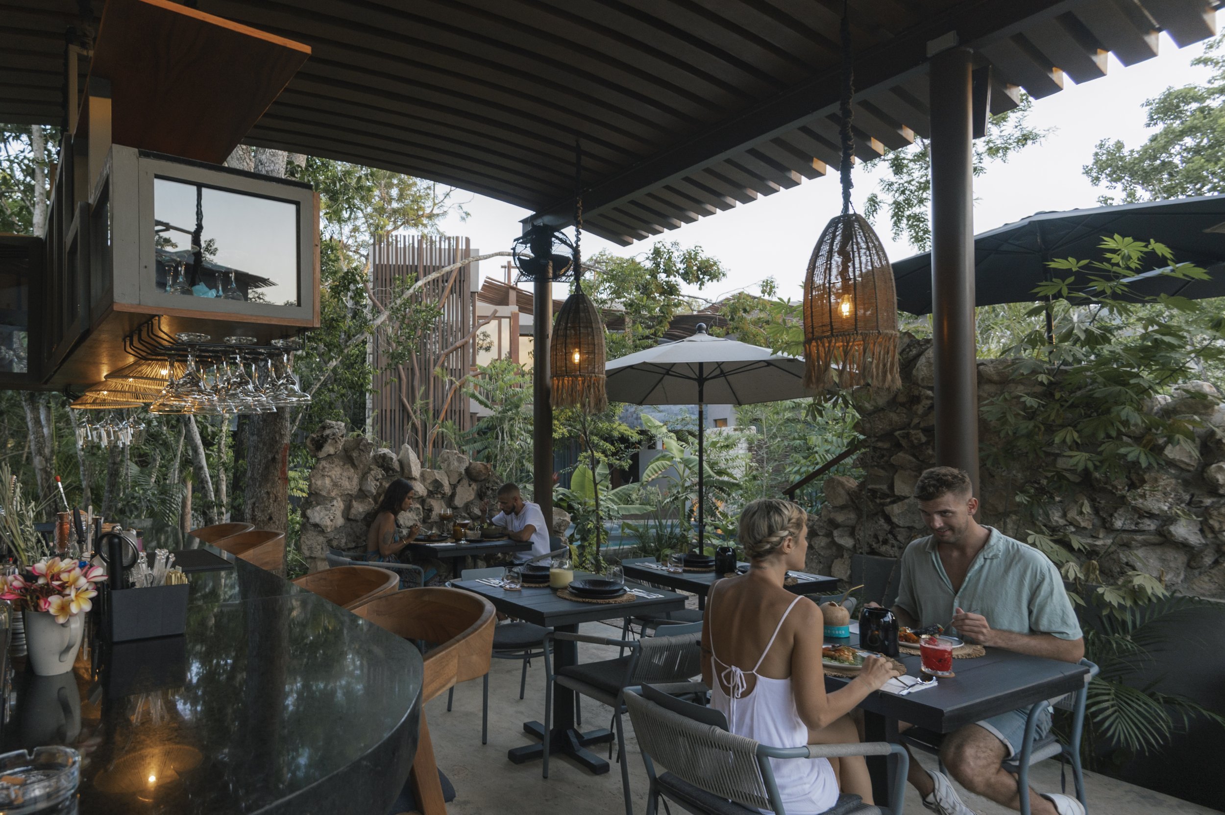 An outdoor restaurant patio with several people dining, surrounded by lush greenery, trees, umbrellas, and hanging lanterns during the evening, at Siwa Tulum.