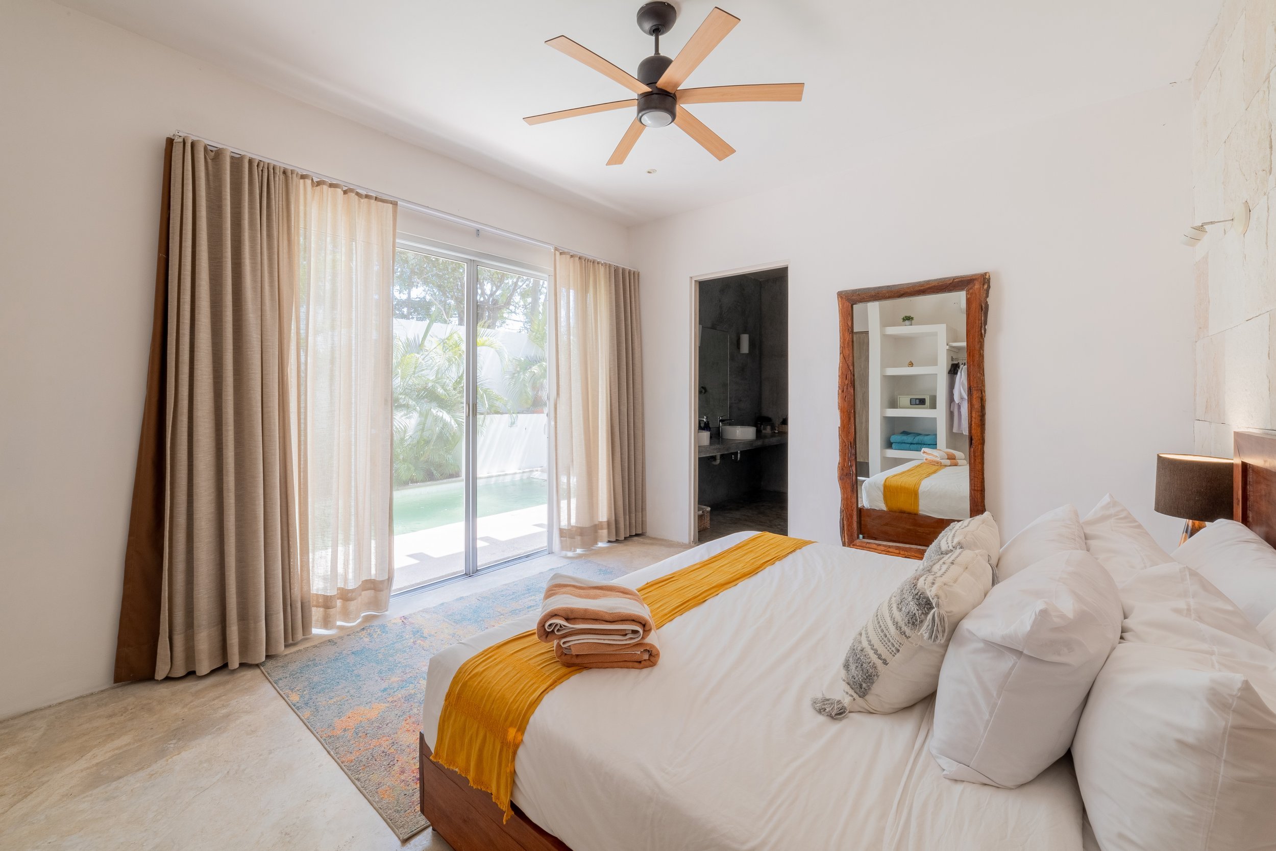 Real estate photography of a bedroom with a bed dressed with white linens and yellow accents, towels on the bed, a large window with beige curtains, at Tulum.
