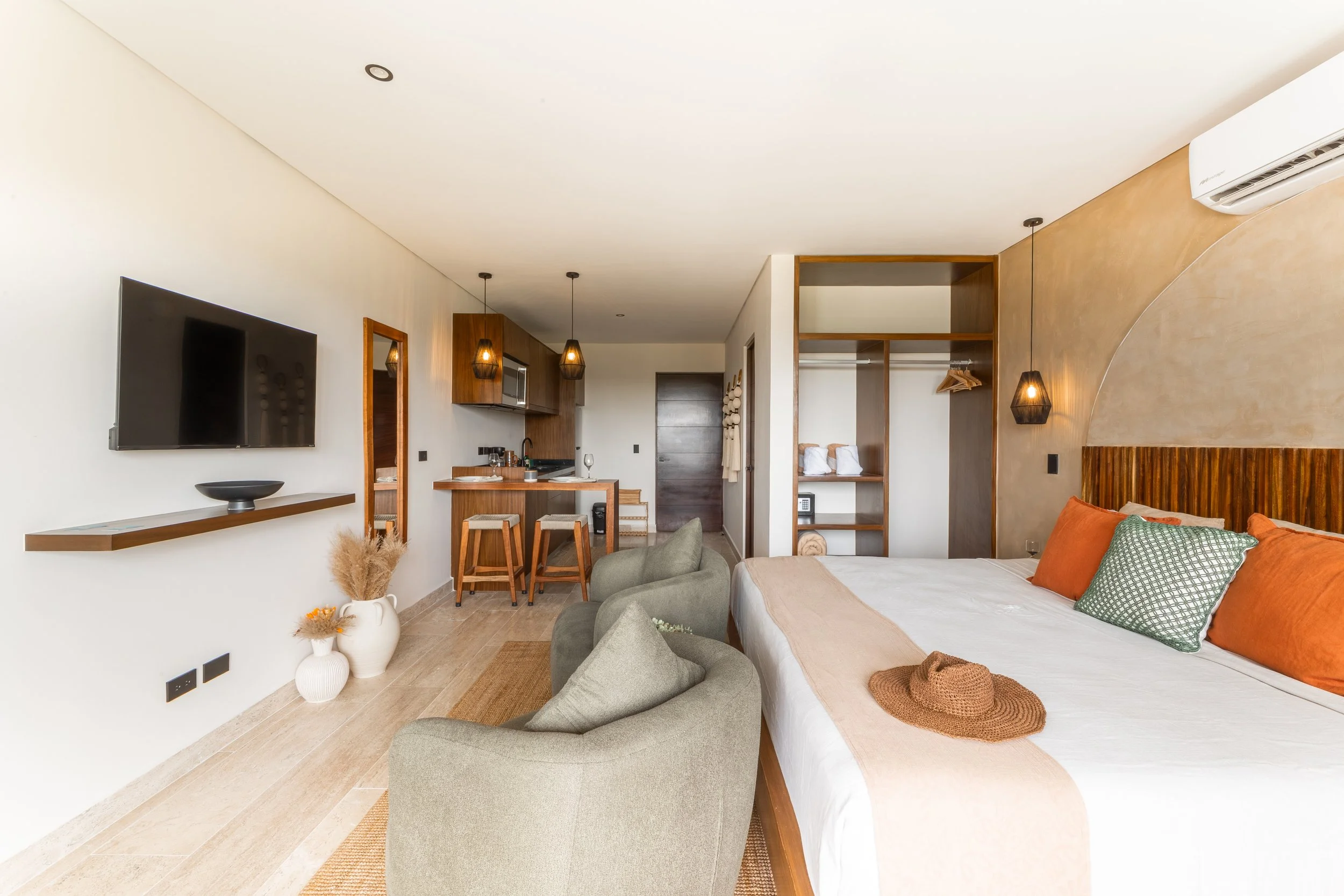 Real estate photography of interior of a modern, stylish hotel room with a bed, orange pillows, a green armchair, a wall-mounted flat-screen TV, and a small dining area with wooden stools and pendant lighting, at mahaual.