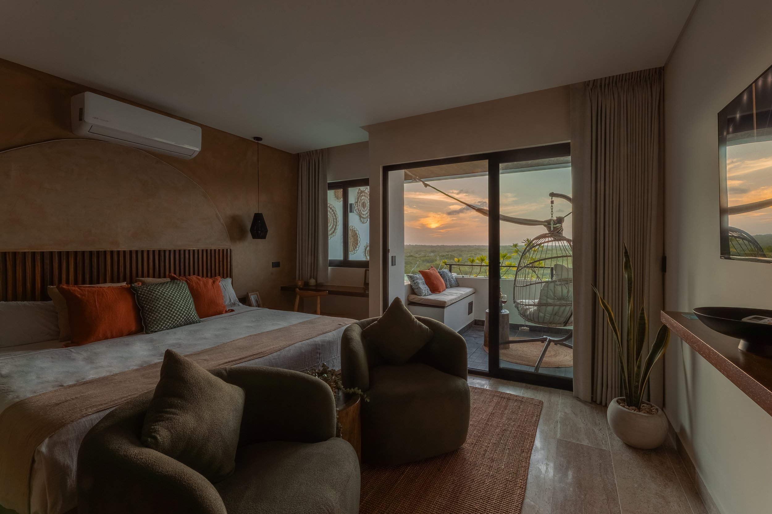 Real estate photography of a cozy bedroom with a large bed decorated with orange and patterned pillows, a sitting area with dark green armchairs, and large windows showing a sunset over greenery with a balcony hanging chair outside, at Mahahual.
