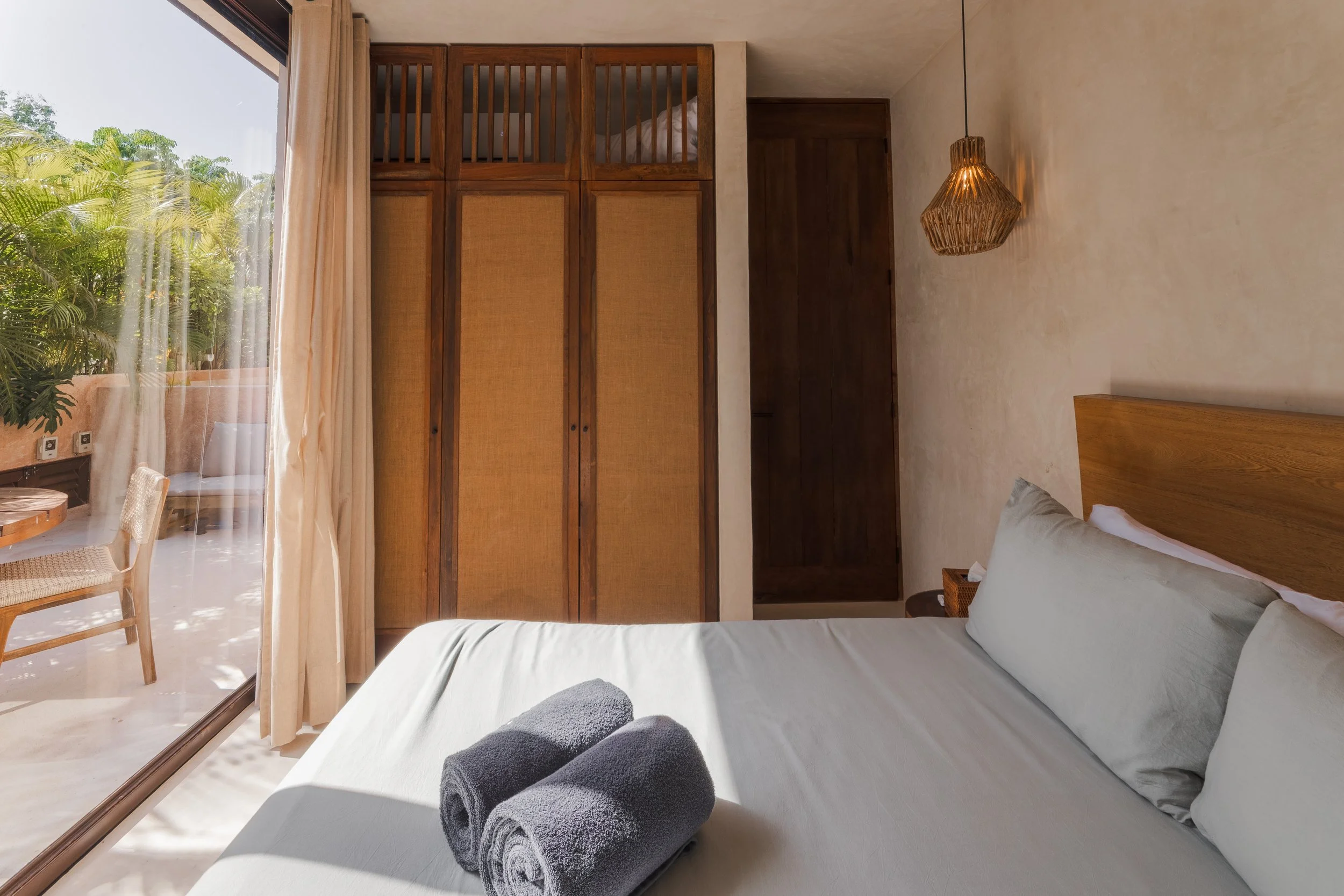 Real estate photography of a bedroom with a bed, pillows, and folded towels, with large glass sliding doors showing a balcony with outdoor furniture and lush greenery outside, at Tulum.