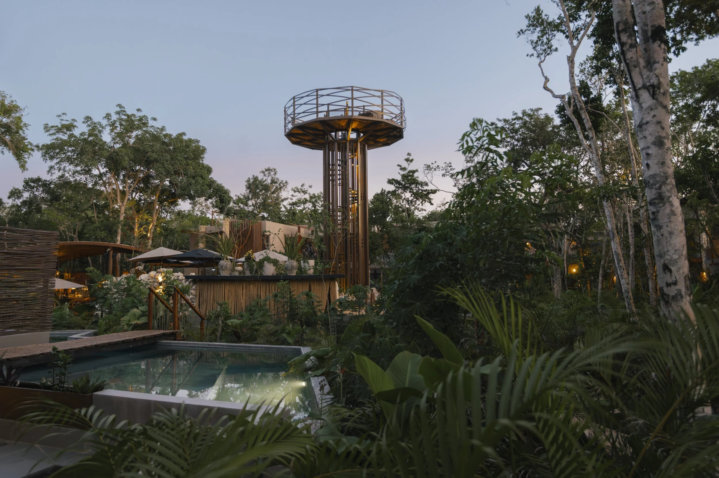 View of an outdoor area with trees, a tower structure, umbrellas, seating areas, and a swimming pool surrounded by lush greenery at dusk, at Siwa Tulum.