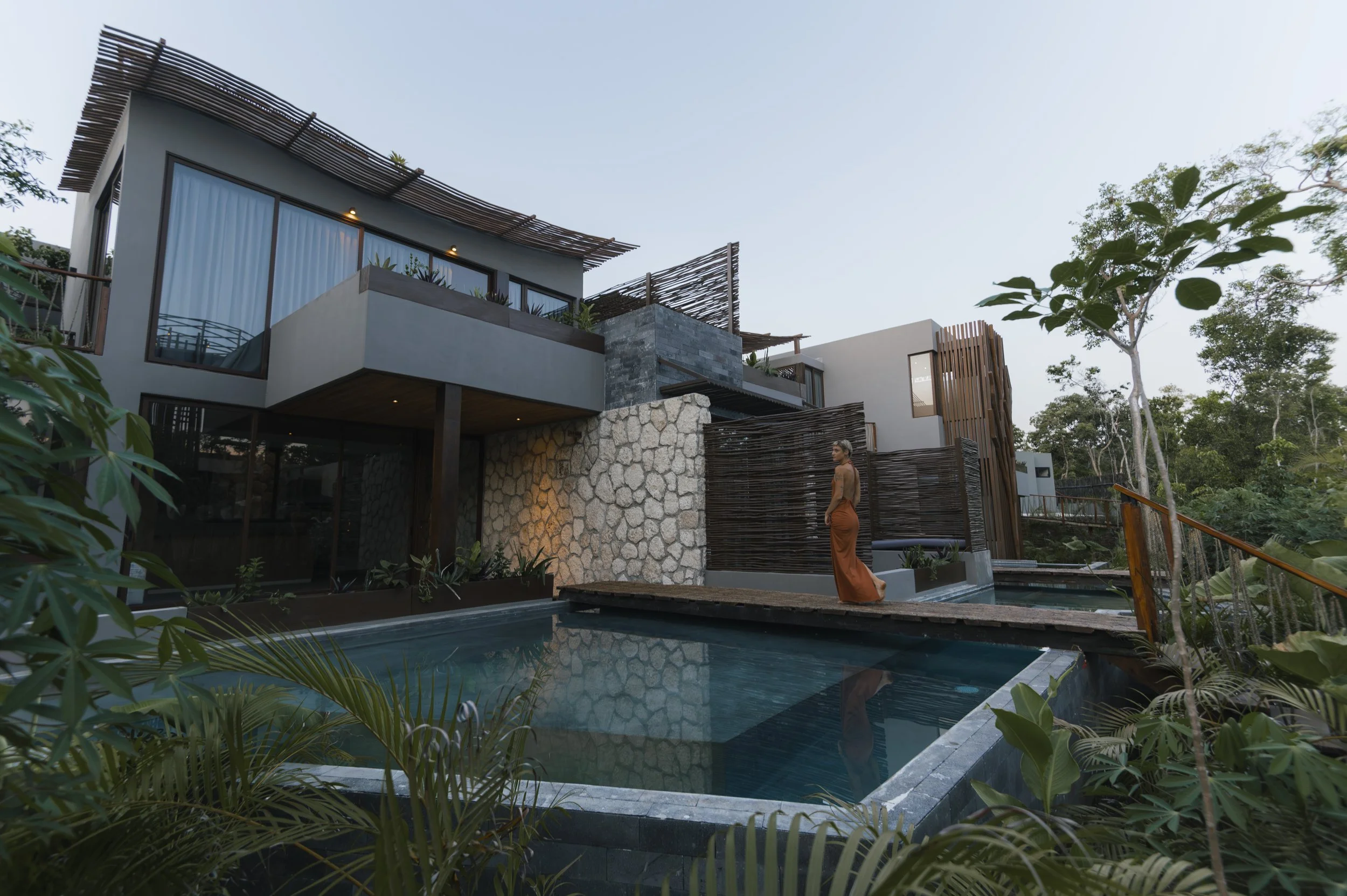 Exterior view of a modern house with large windows, stone and wood accents, and lush greenery around a swimming pool; a woman in a long orange dress standing near the pool, looking to the side, at Siwa Tulum.