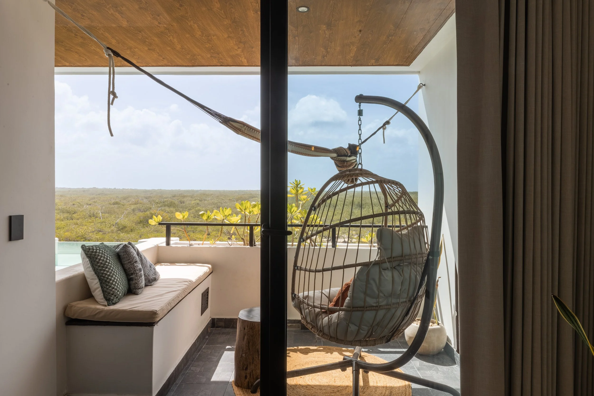 Real estate photography of view of a balcony with outdoor seating, including a cushioned bench with pillows and a hanging wicker chair, overlooking a landscape of greenery under a partly cloudy sky, at Mahahual.