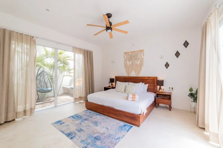Real estate photography of bright bedroom with a wooden bed, white bedding, beige curtains, and a sliding glass door leading to an outside patio with greenery. Decor includes wall hangings and a potted plant, at Tulum.