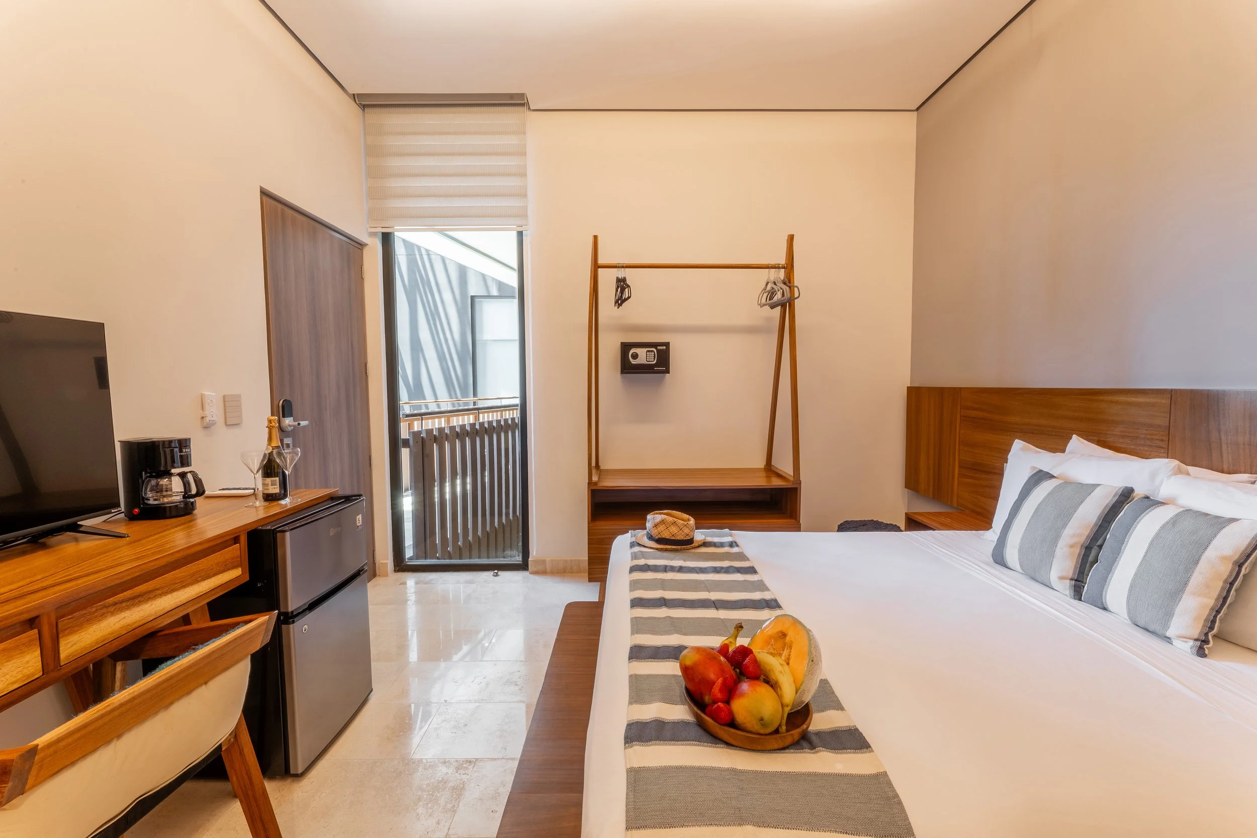 Real estate photography of hotel room with a bed, a wooden headboard, a small table with a fruit basket, a wooden wardrobe, a mini fridge, a coffee maker, a flat-screen TV, a balcony door with a roller shade, and a clothing rack, at Amira Tulum.