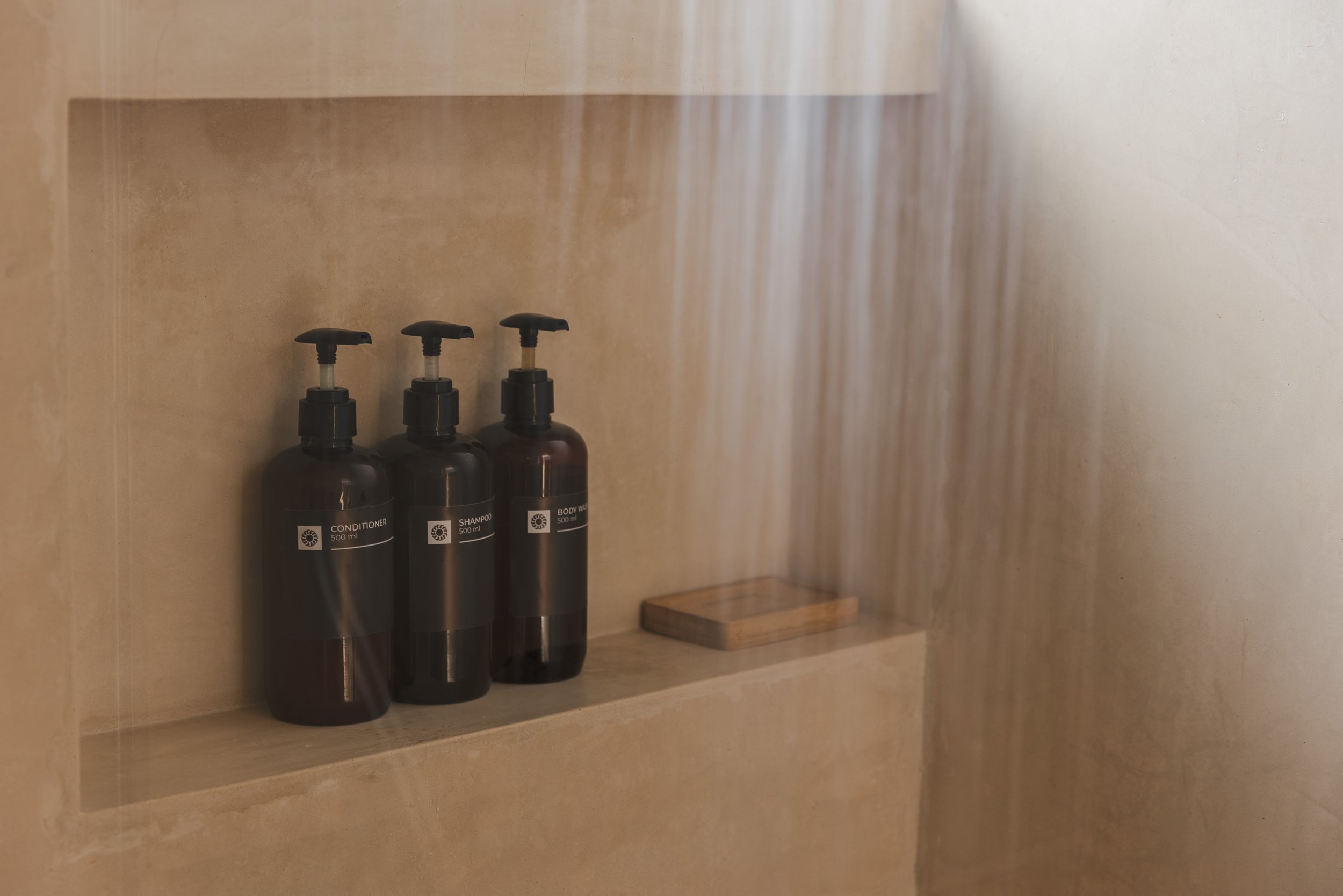 Real estate photography of three black pump bottles of conditioner, shampoo, and body wash on a beige bathroom shelf, with a small wooden platform next to them, at Tulum.