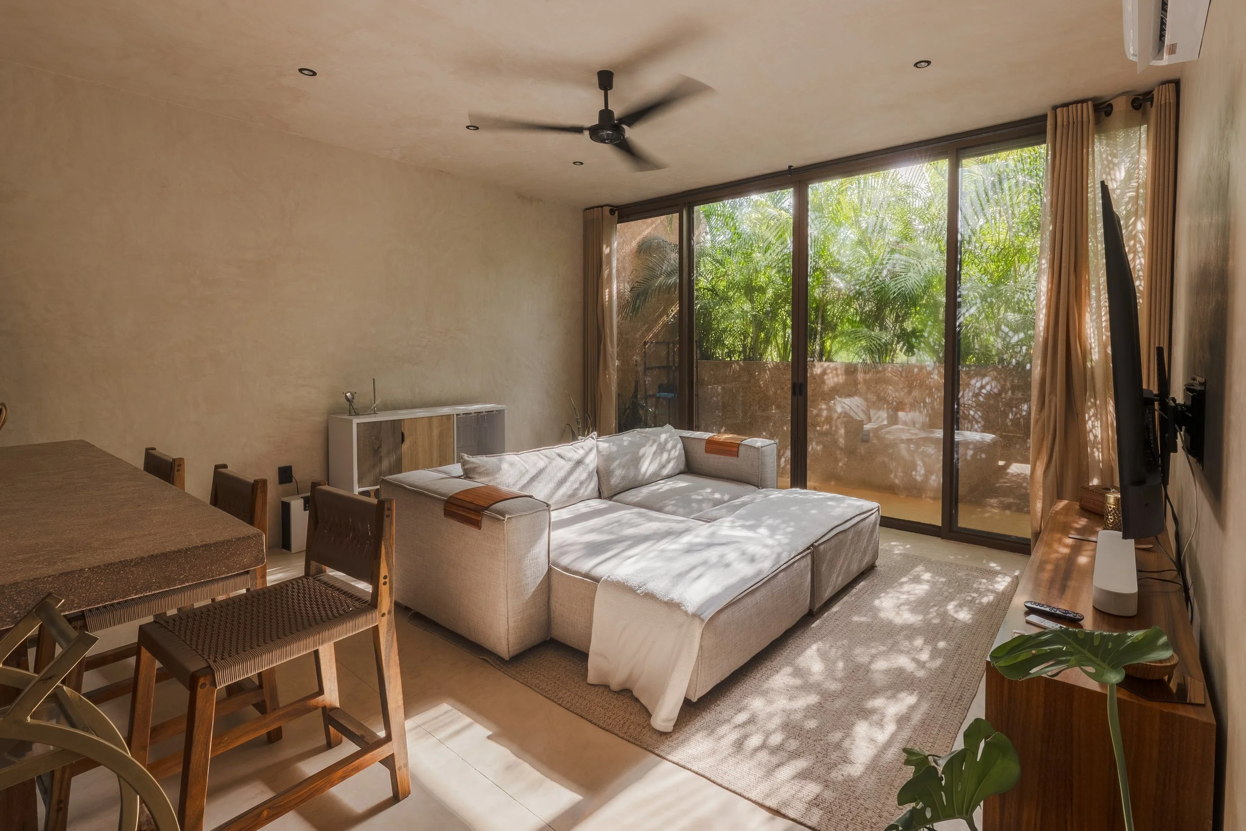 Real estate photography of living room with beige walls, sunlight streaming through large sliding glass doors, cream sofa, wooden dining table with chairs, ceiling fan, and green plants at Tulum