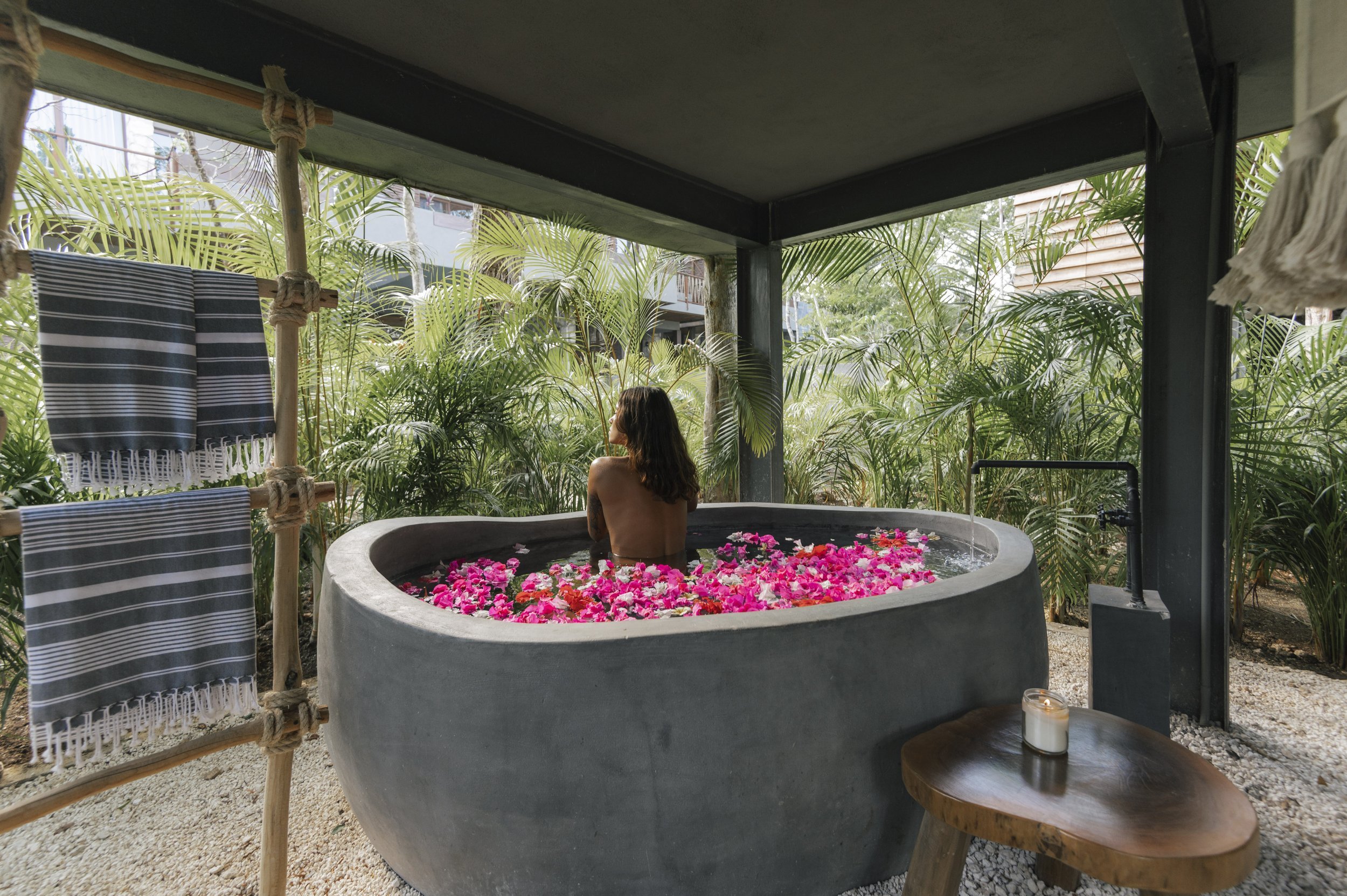 A woman relaxing in a round concrete bathtub filled with pink and white flower petals, surrounded by lush green tropical plants, under a shaded outdoor structure with hanging towels and a small wooden side table with a white candle, at Siwa Tulum.