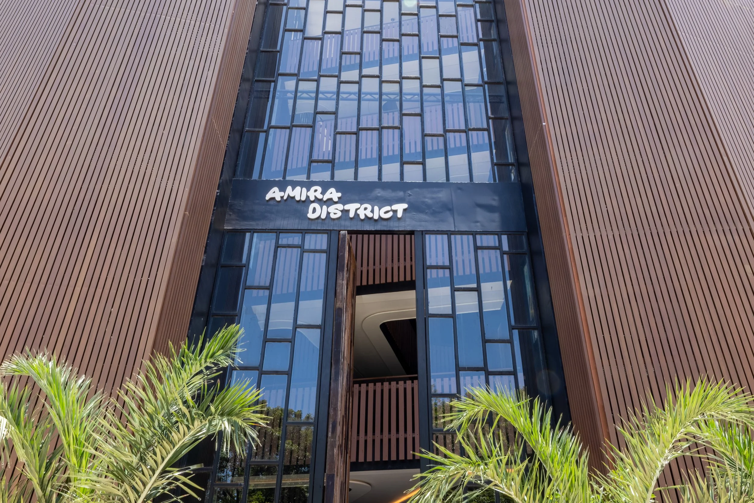 Real estate photography of tall modern building with wooden slat exterior, large glass windows, and a sign that reads 'Amira District' at the entrance. 