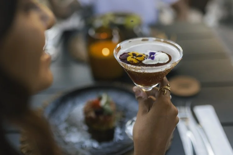 Person holding a cocktail glass with flowers, sitting at a table with a plate of food, utensils, and another blurred person in the background, at Siwa Tulum.