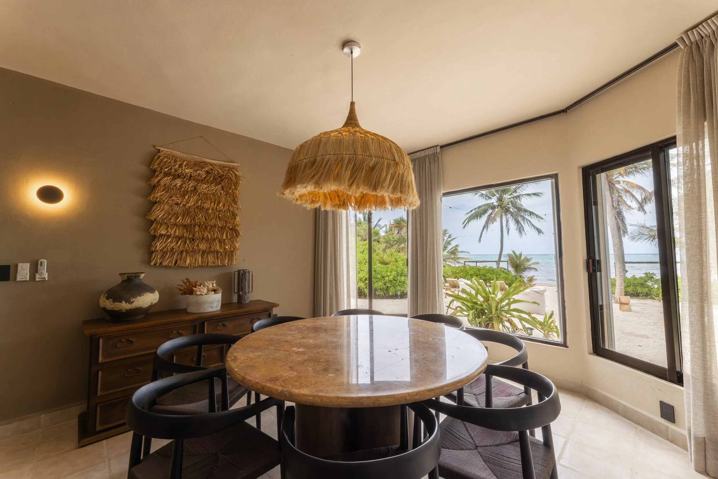 Interior of a room with a round marble dining table surrounded by six black chairs. A large, woven, fringed pendant light hangs above the table, at Bespoke Tulum.