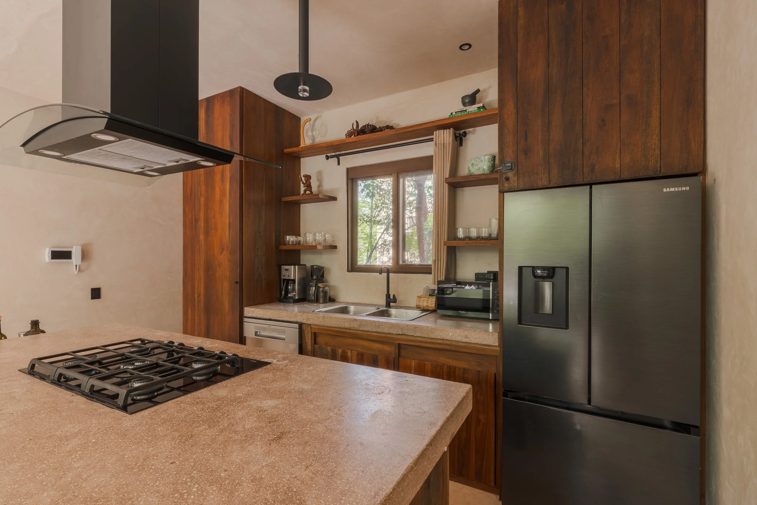 Real estate photography of a kitchen with wooden cabinets, a large window, a countertop with a stovetop, a coffee maker, a microwave, and a refrigerator, at Tulum.