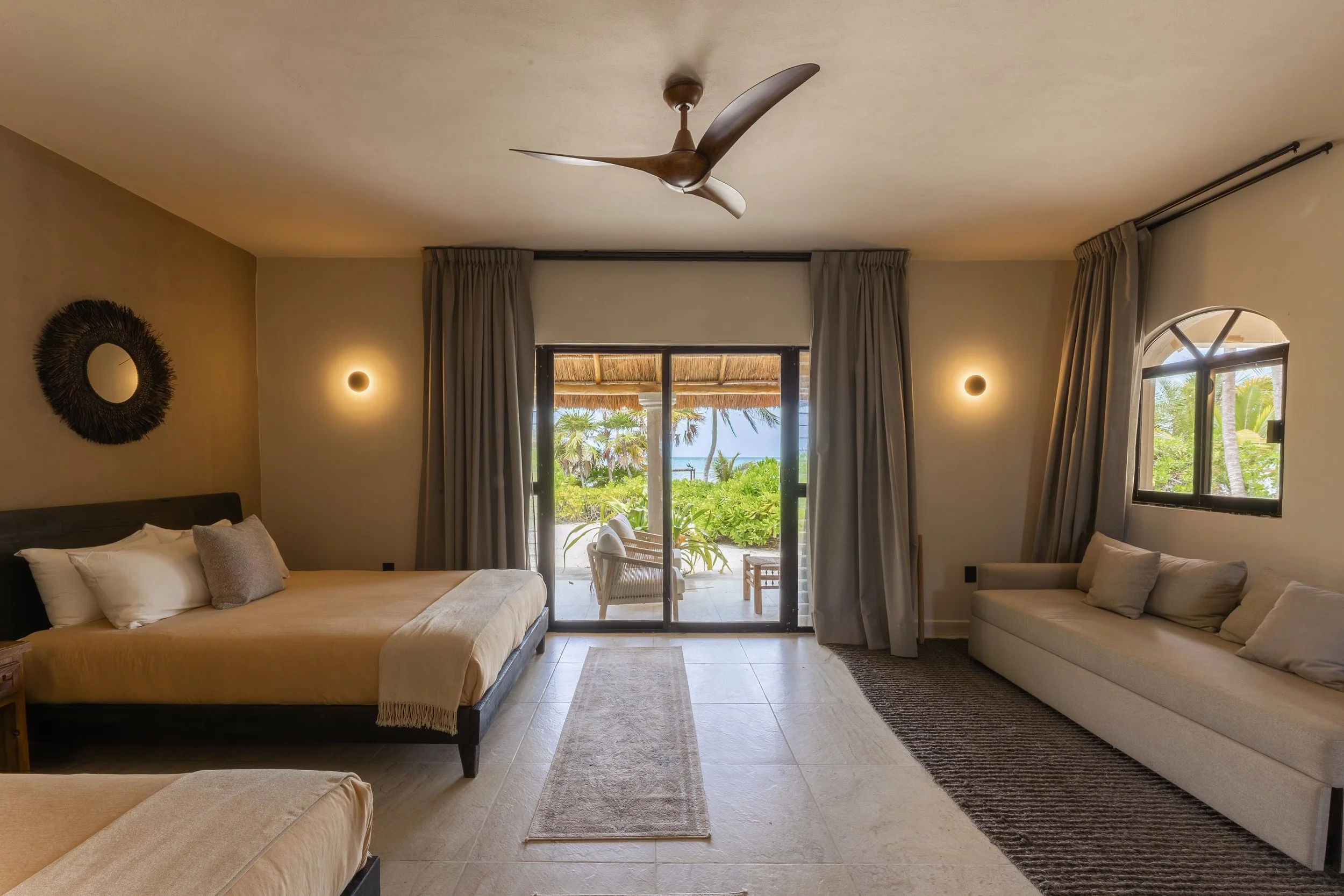 A hotel room with a large bed, a sofa, sliding glass door with a view of tropical plants and the ocean, ceiling fan, and modern wall lighting, at Bespoke Tulum.