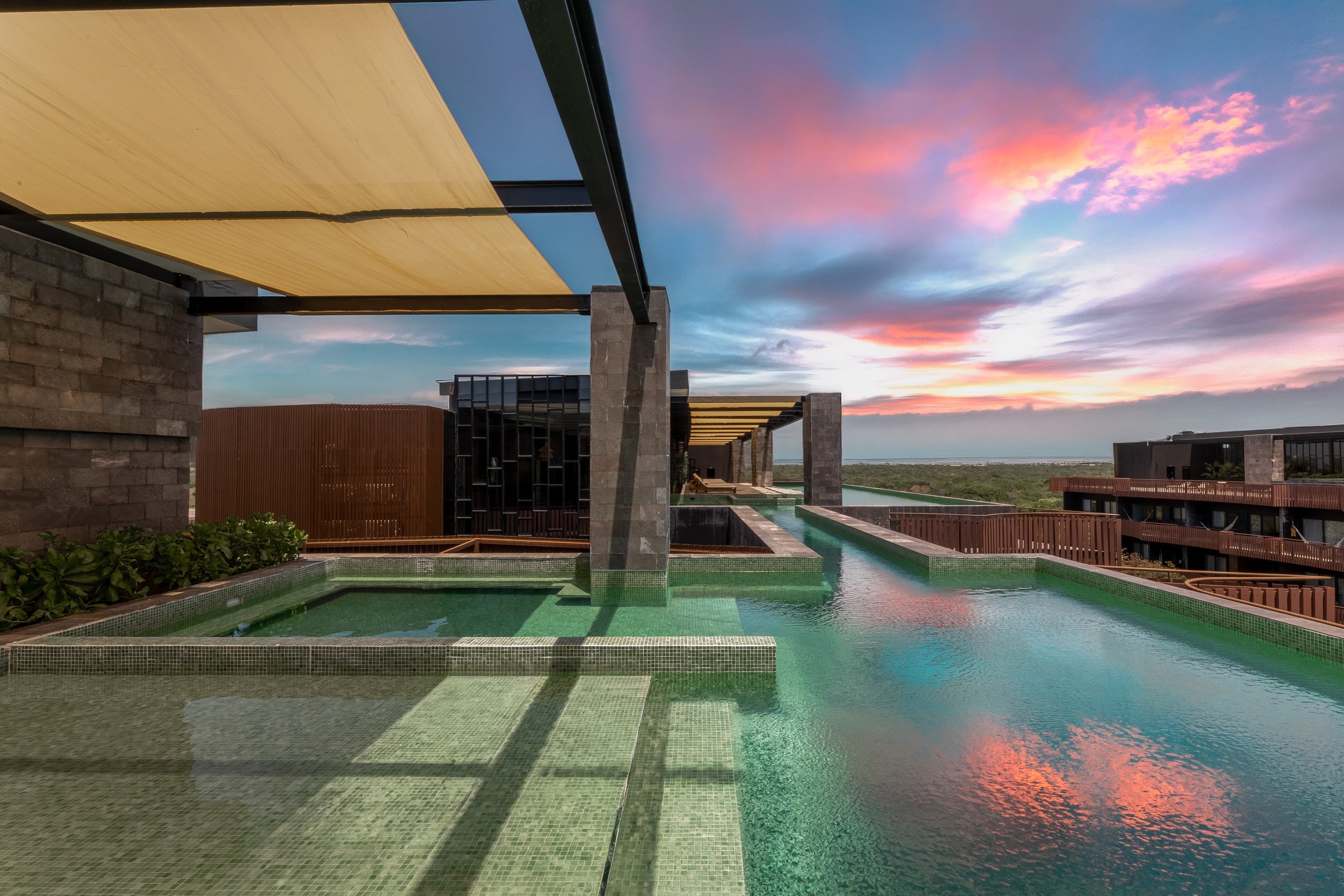 Real estate photography of an  outdoor swimming pool with clear green water, surrounded by a modern building with stone and wood accents, and a colorful sky with pink and purple clouds at sunset, at Amira Tulum.