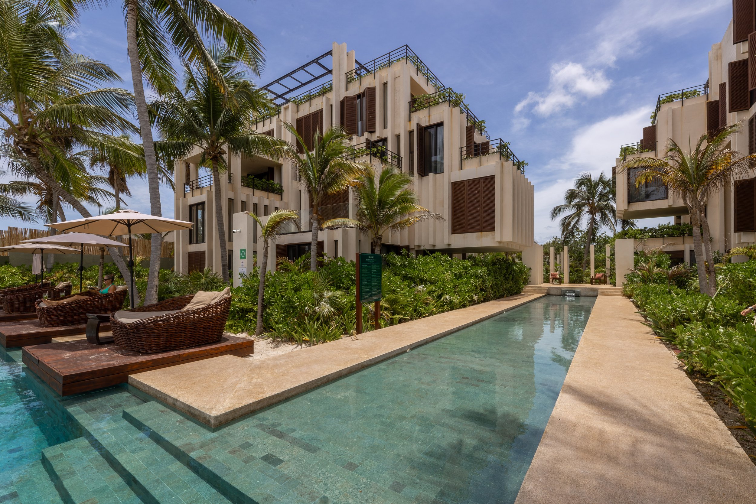 Luxury hotel with a swimming pool surrounded by tropical palm trees and modern white buildings with brown accents, umbrellas, and outdoor seating, at Bespoke Tulum.
