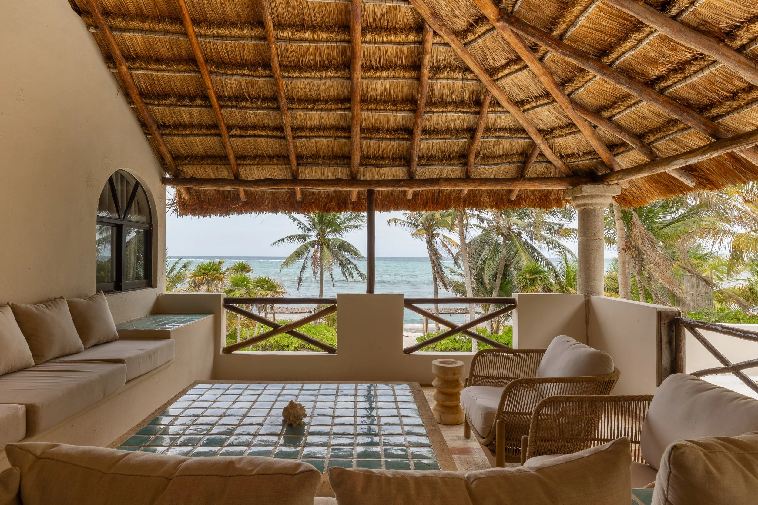 Beachside patio with white cushioned furniture, tiled table, and thatched roof, overlooking palm trees and the ocean, at Bespoke Tulum.