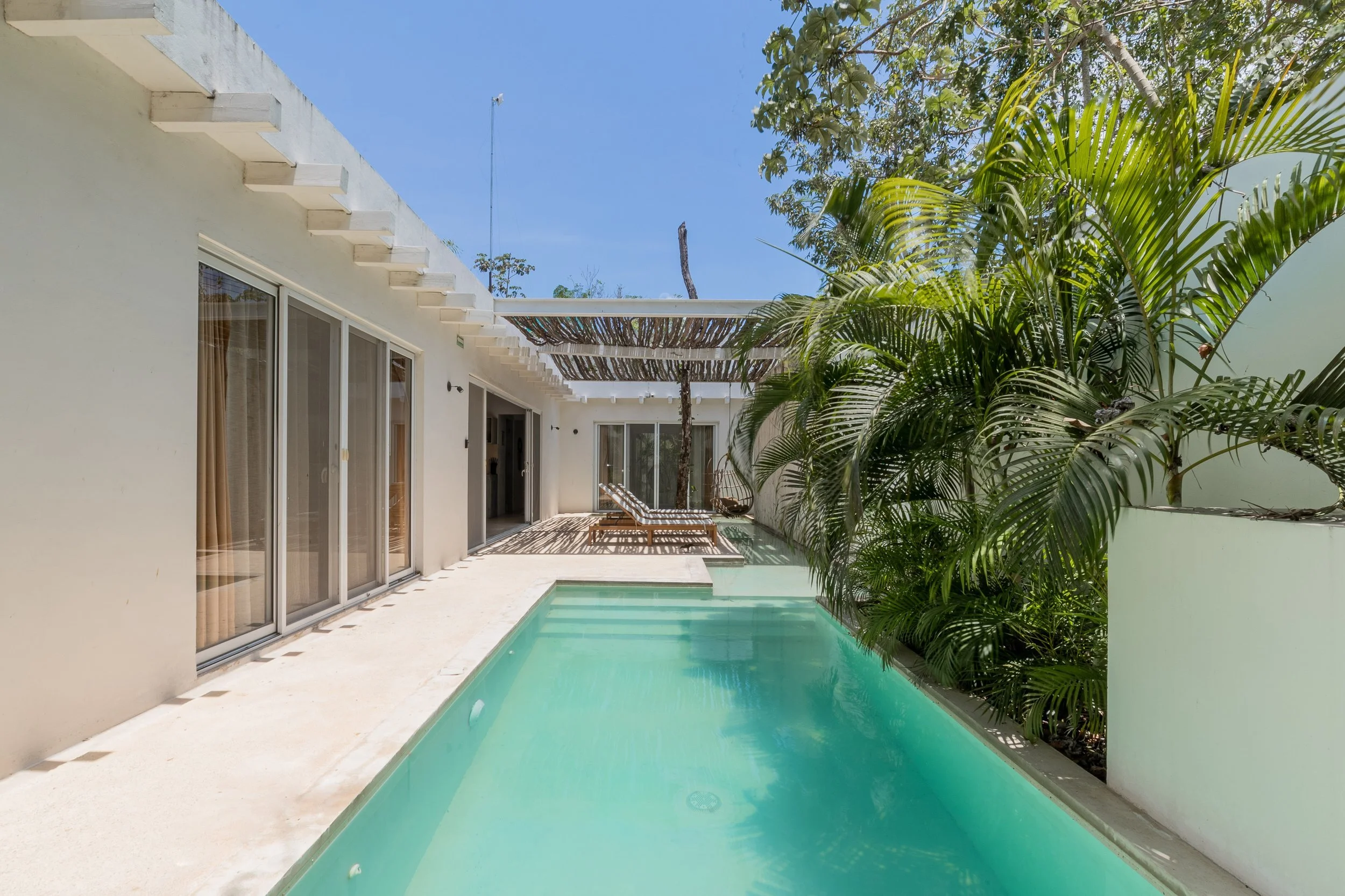 Real estate photography of a modern house with sliding glass doors leading to a outdoor swimming pool, surrounded by green palm trees and a clear blue sky, at Tulum.