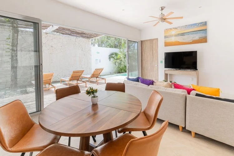 Real estate photography of living room with a large sliding glass door leading to a patio with four wooden lounge chairs, at Tulum.