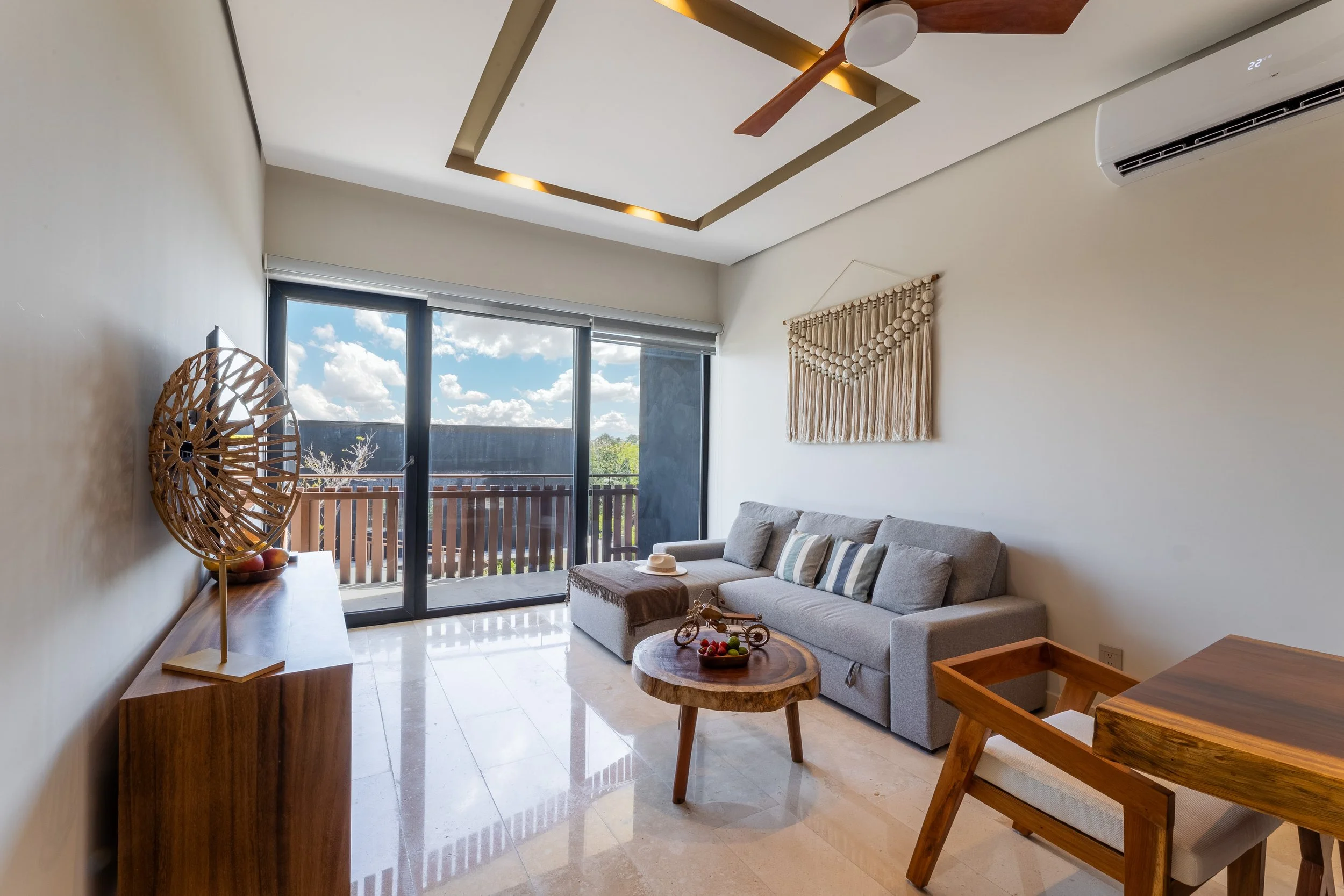Real estate photography of sunlit living room with beige walls, a large window with a balcony, beige sofa with pillows, at Amira Tulum