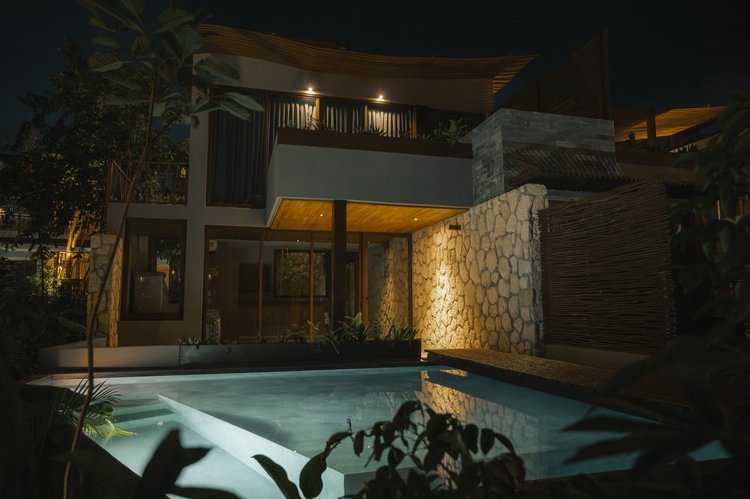 Nighttime view of a modern house with a swimming pool in the foreground, illuminated exterior lighting, stone and wood exterior walls, large windows, and lush plants surrounding the area, at Siwa Tulum.