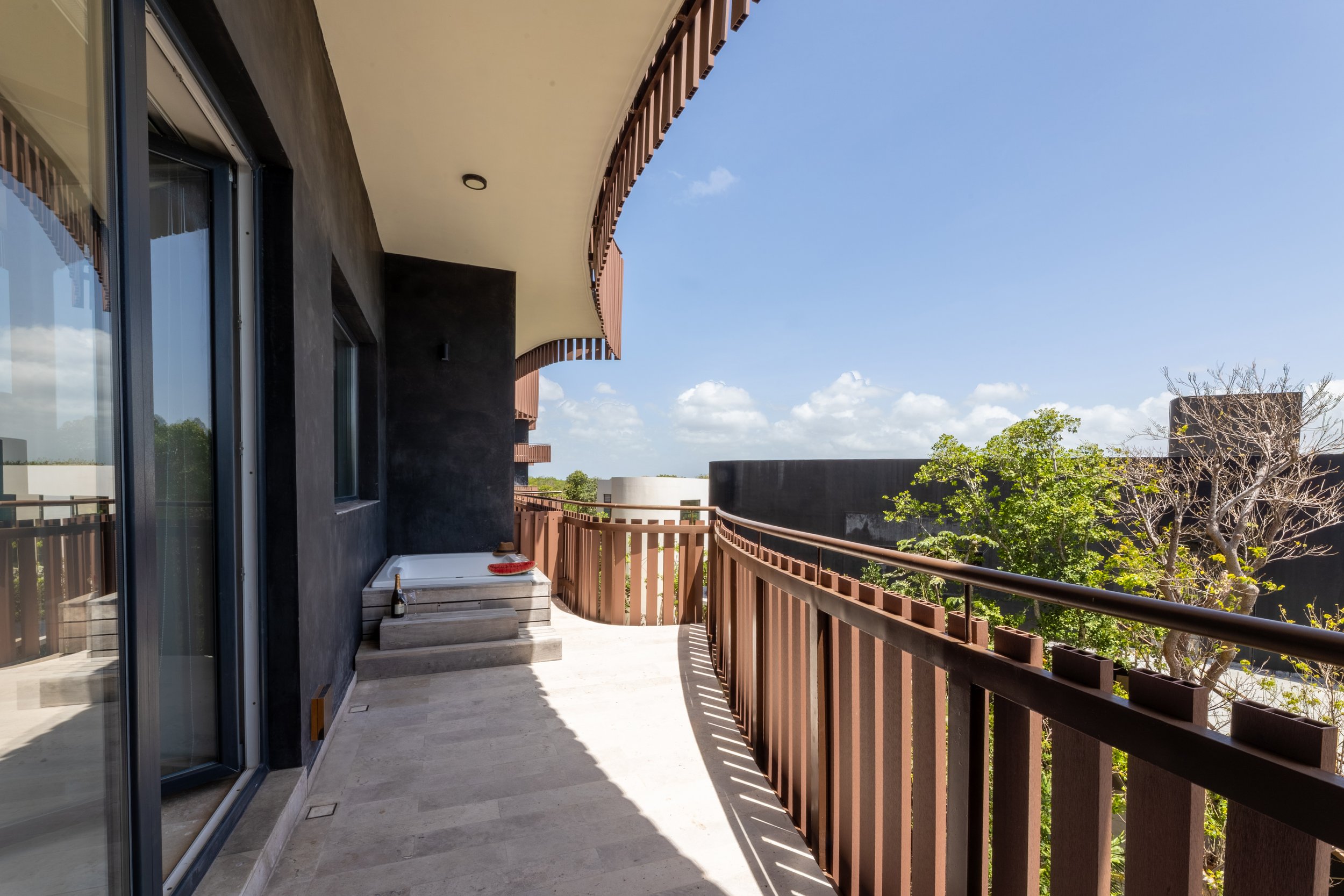 Real estate photography of balcony with sliding glass doors, wooden railing, a small table with a bottle and a red plate, and a view of trees and blue sky, at Amira Tulum