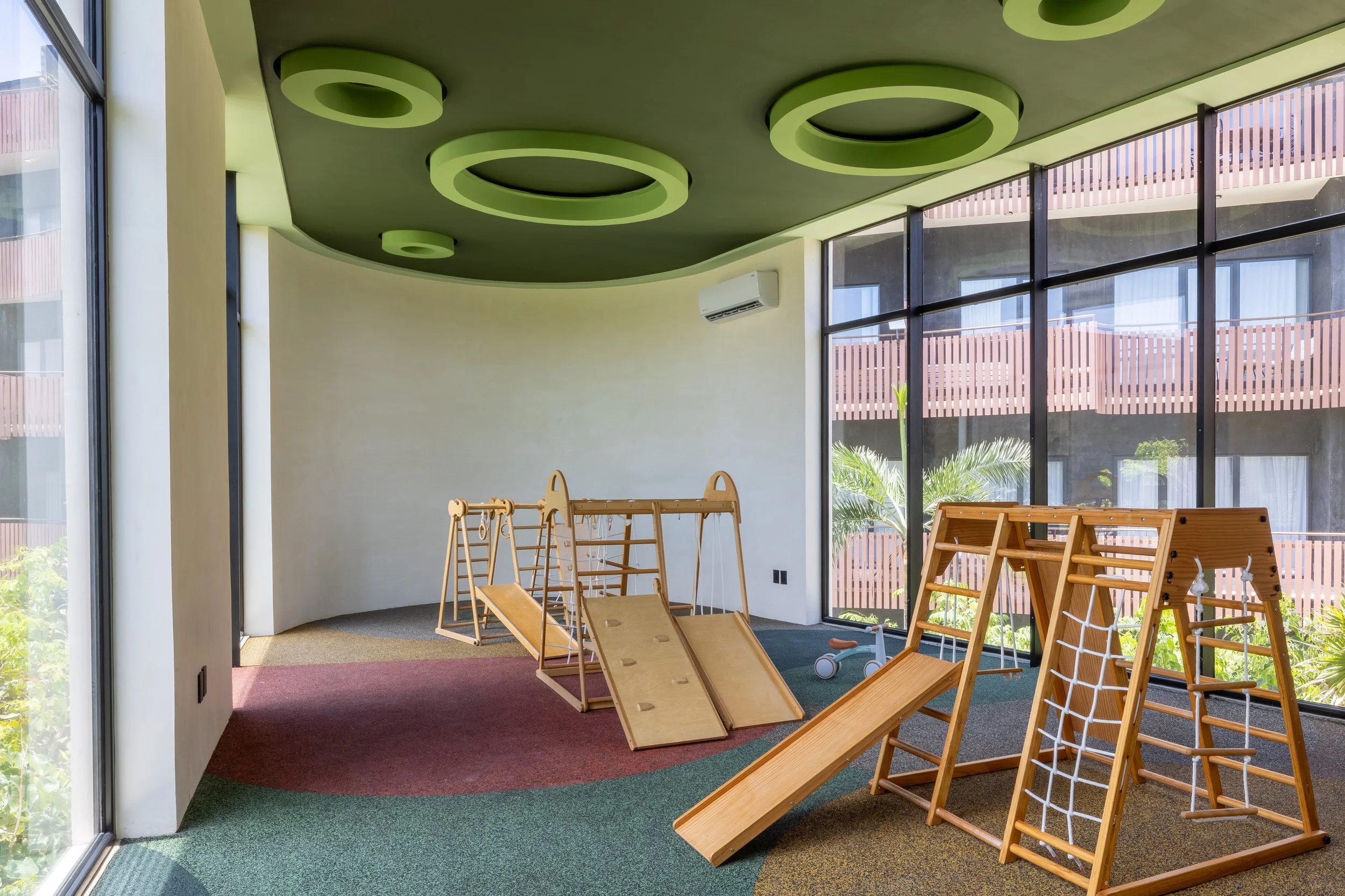 Real estate photography of indoor play area with wooden climbing structures, slides, and mats, next to floor-to-ceiling windows showing outdoor buildings and greenery, at Amira Tulum.