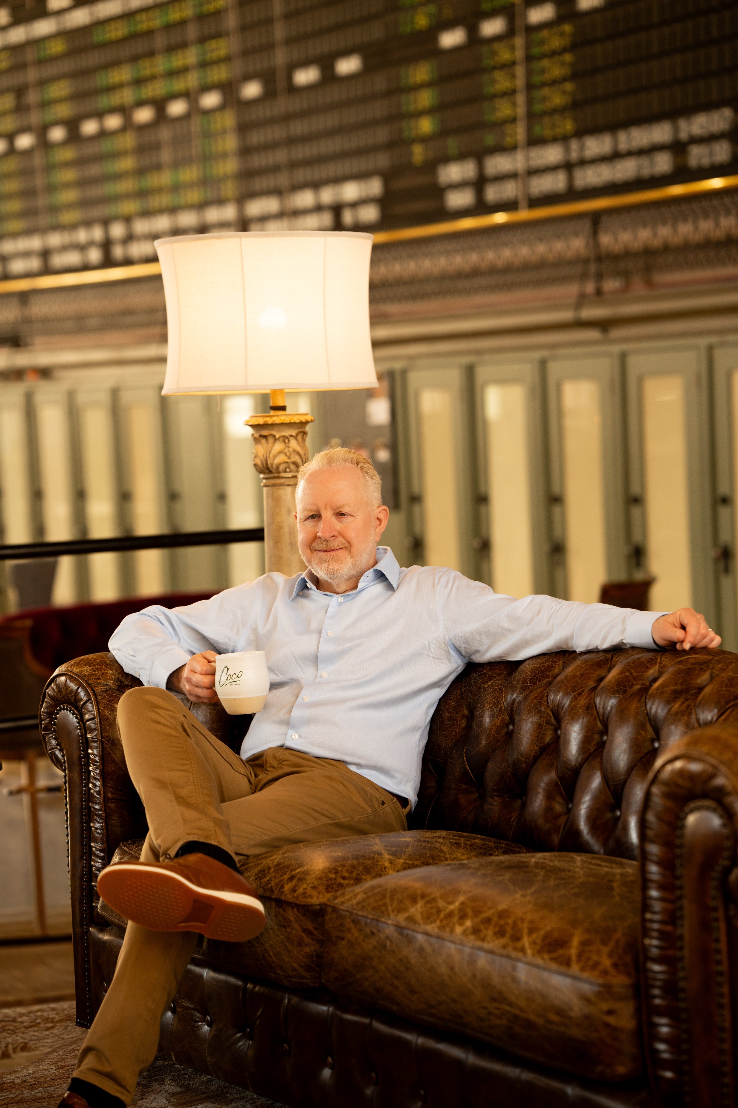 A man with gray hair and a beard sitting on a vintage brown leather couch in an airport lounge, holding a white mug, with a large illuminated lampshade behind him and rows of luggage storage lockers in the background.