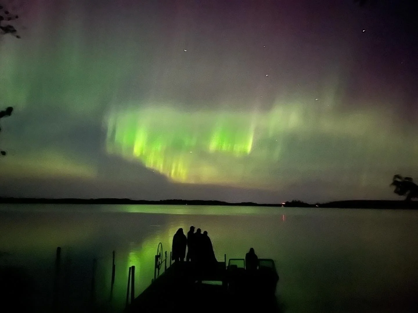 Nighttime scene of a Minnesotan aurora borealis with green and purple hues over a calm lake, with a small group of people standing on a dock watching the spectacle.