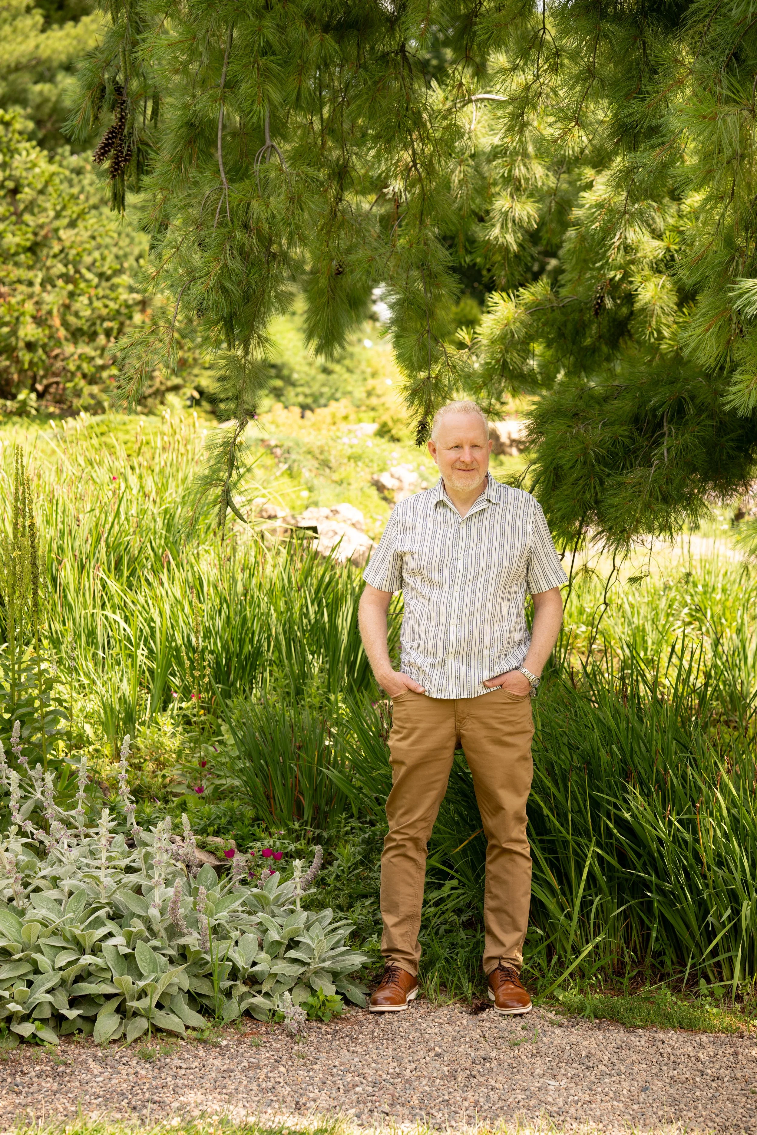 A man standing outdoors surrounded by lush green plants, trees, and flowers on a gravel path.
