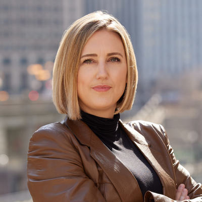 A woman with shoulder-length blonde hair standing outdoors in an urban setting, wearing a brown leather jacket and black turtleneck, with a city skyline in the background.