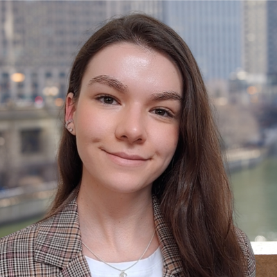 Young woman with long brown hair smiling outdoors in front of a cityscape and river.