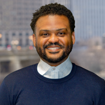 Portrait of a smiling man with curly hair and a beard, wearing a navy blue sweater over a light blue collared shirt, with a cityscape background.