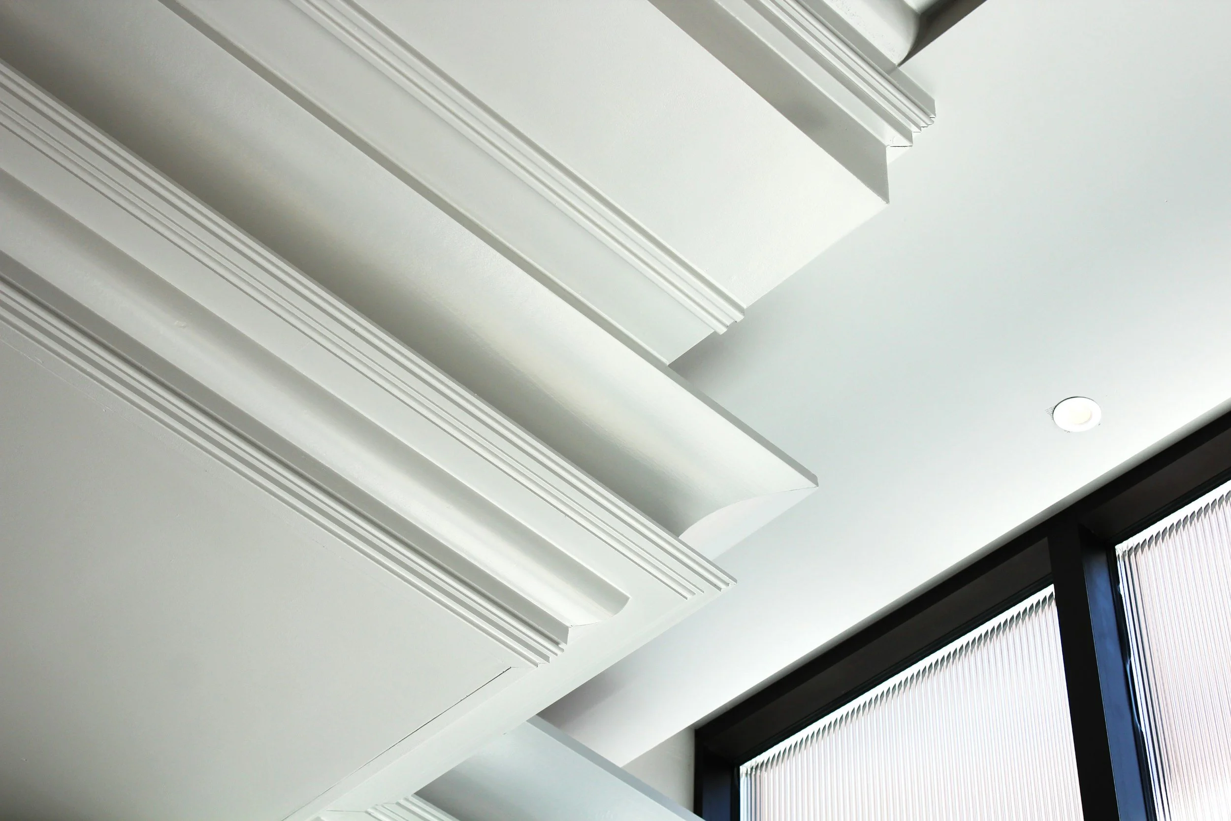 White ceiling with decorative molding and a circular recessed light, adjacent to a window with vertical blinds.