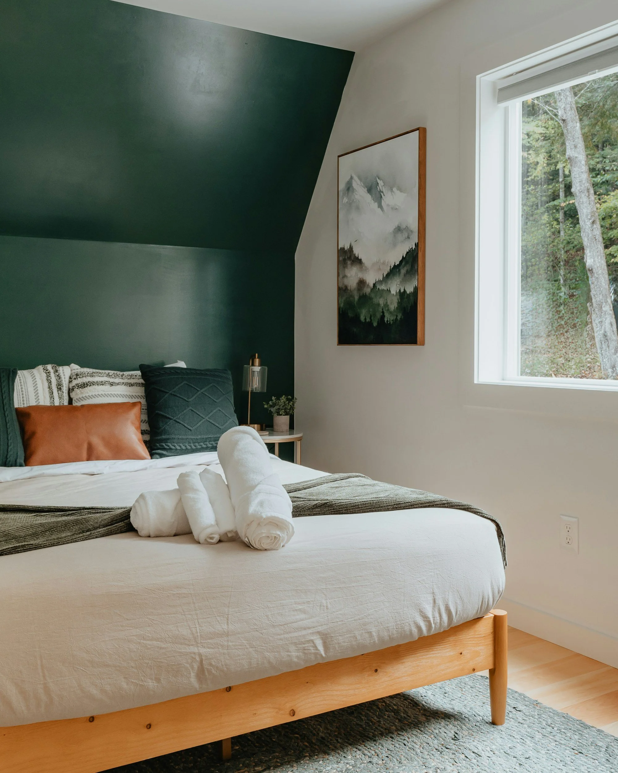 A neatly made bed with white linens, two rolled white towels on top, and multiple pillows including a tan pillow, a dark green quilted pillow, and a black pillow, in a modern bedroom with a green accent wall, mountain landscape artwork, a window with a view of trees, a small bedside table with a plant, and a floor rug.