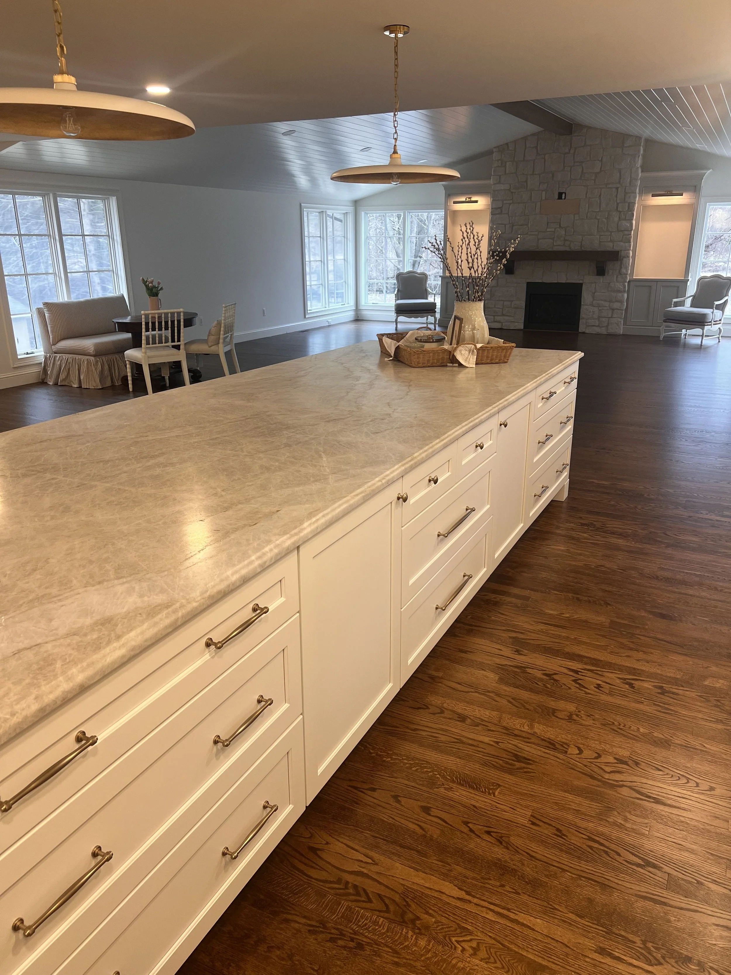 Interior of a living room with a kitchen island in the foreground, featuring a beige marble countertop and white cabinetry with brass handles. The room has large windows, a stone fireplace, and hardwood floors, with some chairs and floral decorations