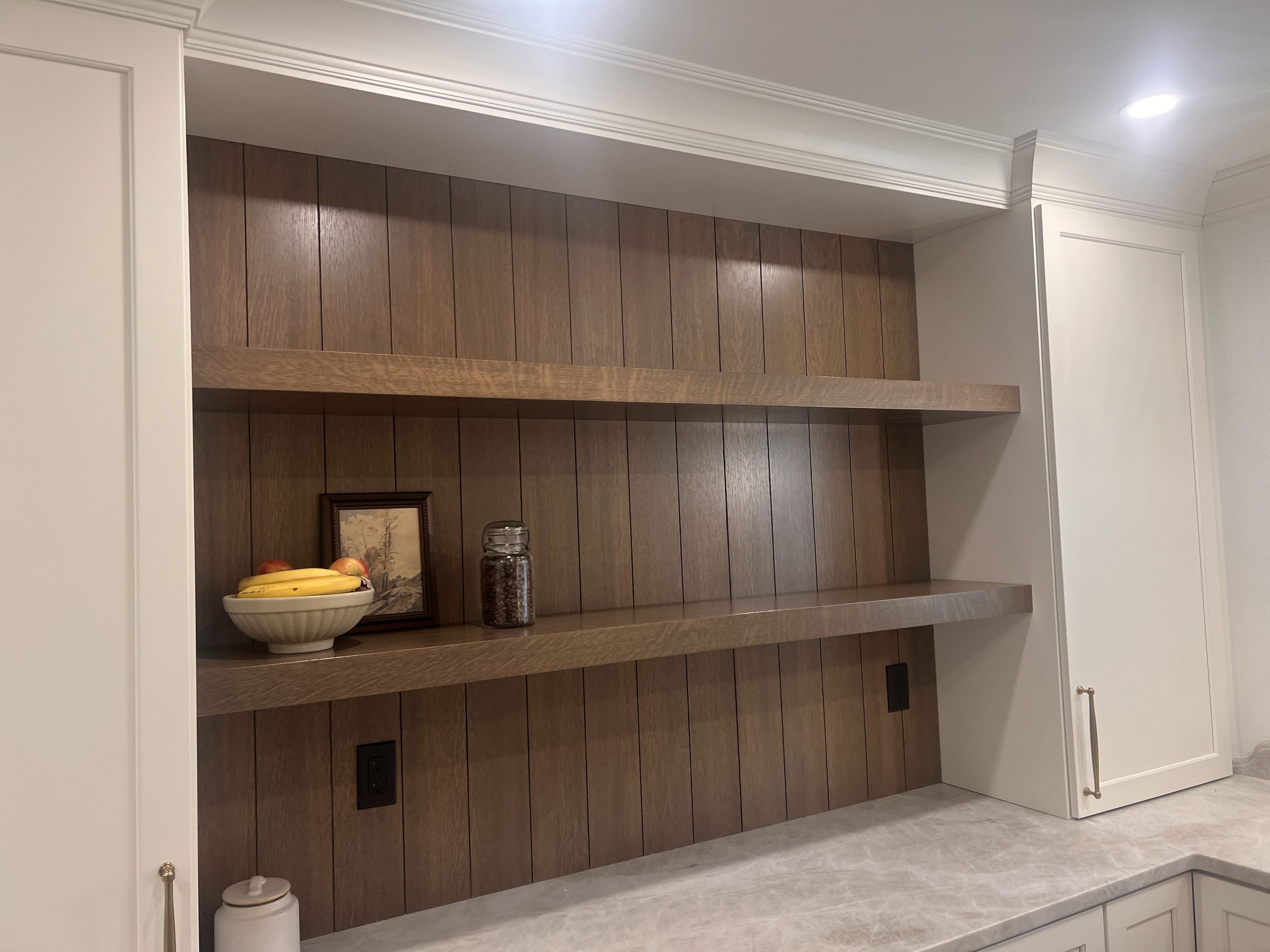 Interior of a kitchen with black electrical outlets, white walls, crown molding, and wooden wall panels. There is a shelf holding a bowl of bananas and apples, and a framed picture along with jars.