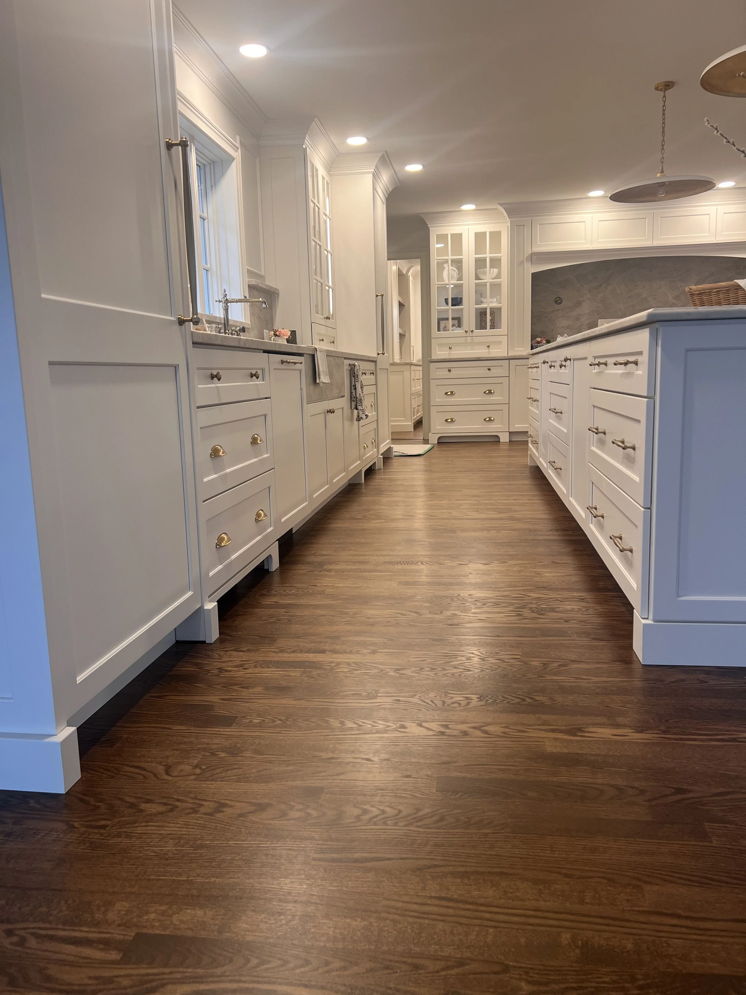 Bright kitchen with white cabinets, wooden flooring, and recessed ceiling lights. Features a large central island with cabinet storage, a sink under a window, and glass-fronted upper cabinets.