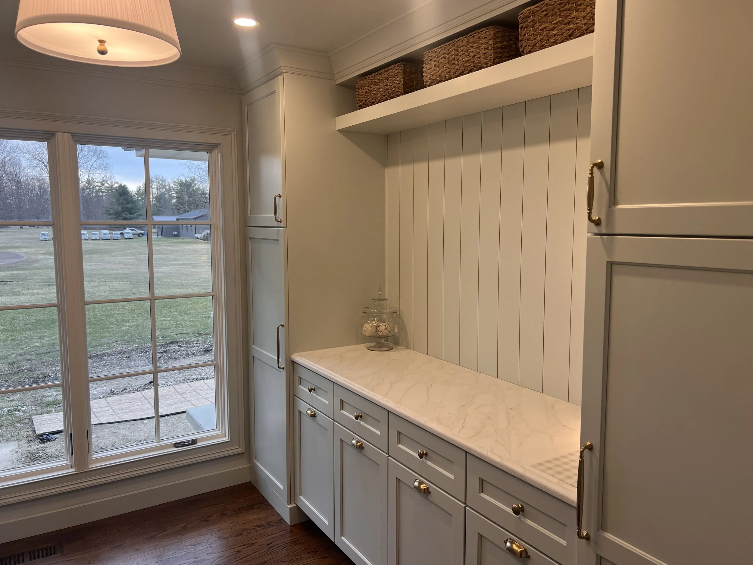 Kitchen with white cabinets, gold handles, marble countertop, and a large window overlooking a yard with trees and parked cars.