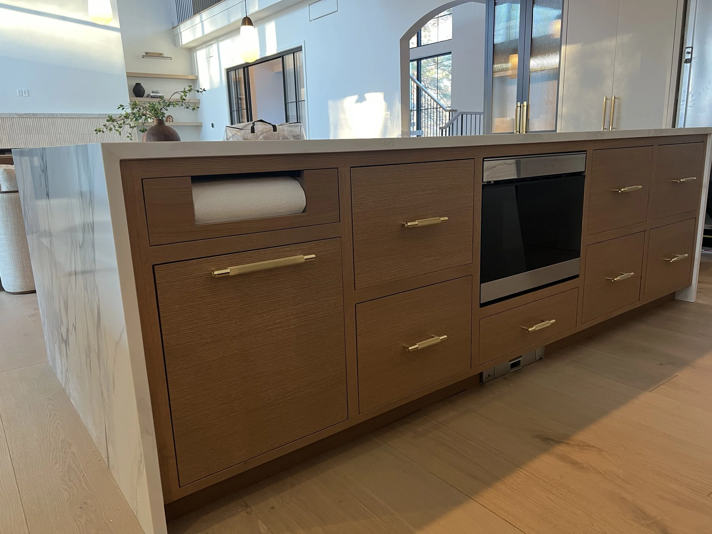 Modern kitchen island with a wooden base, gold handles, and a white marble countertop. A paper towel roll is on a built-in holder on the side. In the background, there are white walls, shelves with decorative items, a potted plant, large windows, and