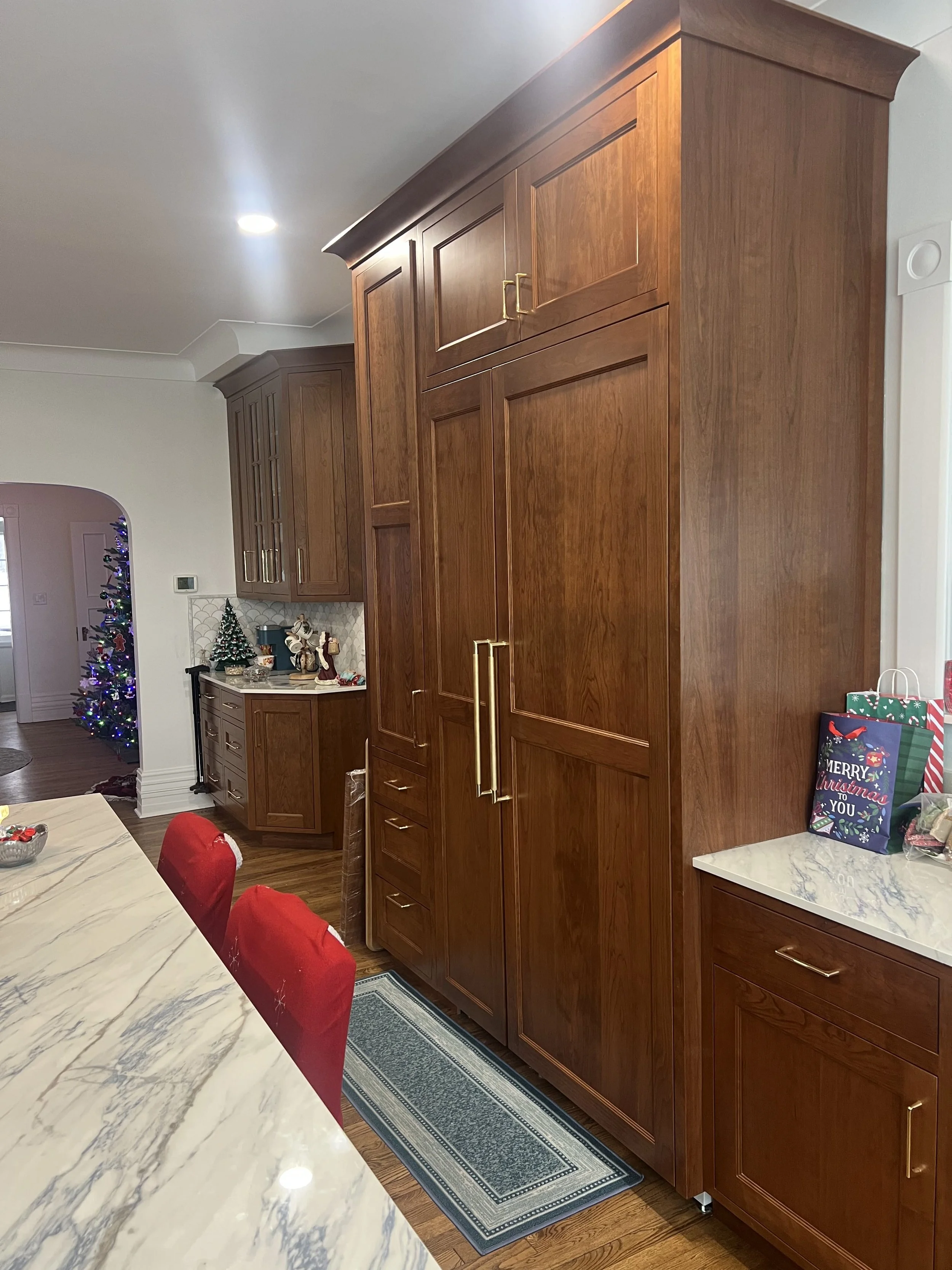 Kitchen with large wooden cabinets, marble countertop, holiday decor including Christmas tree, presents, and a Santa figurine, with a dining table and red chairs.