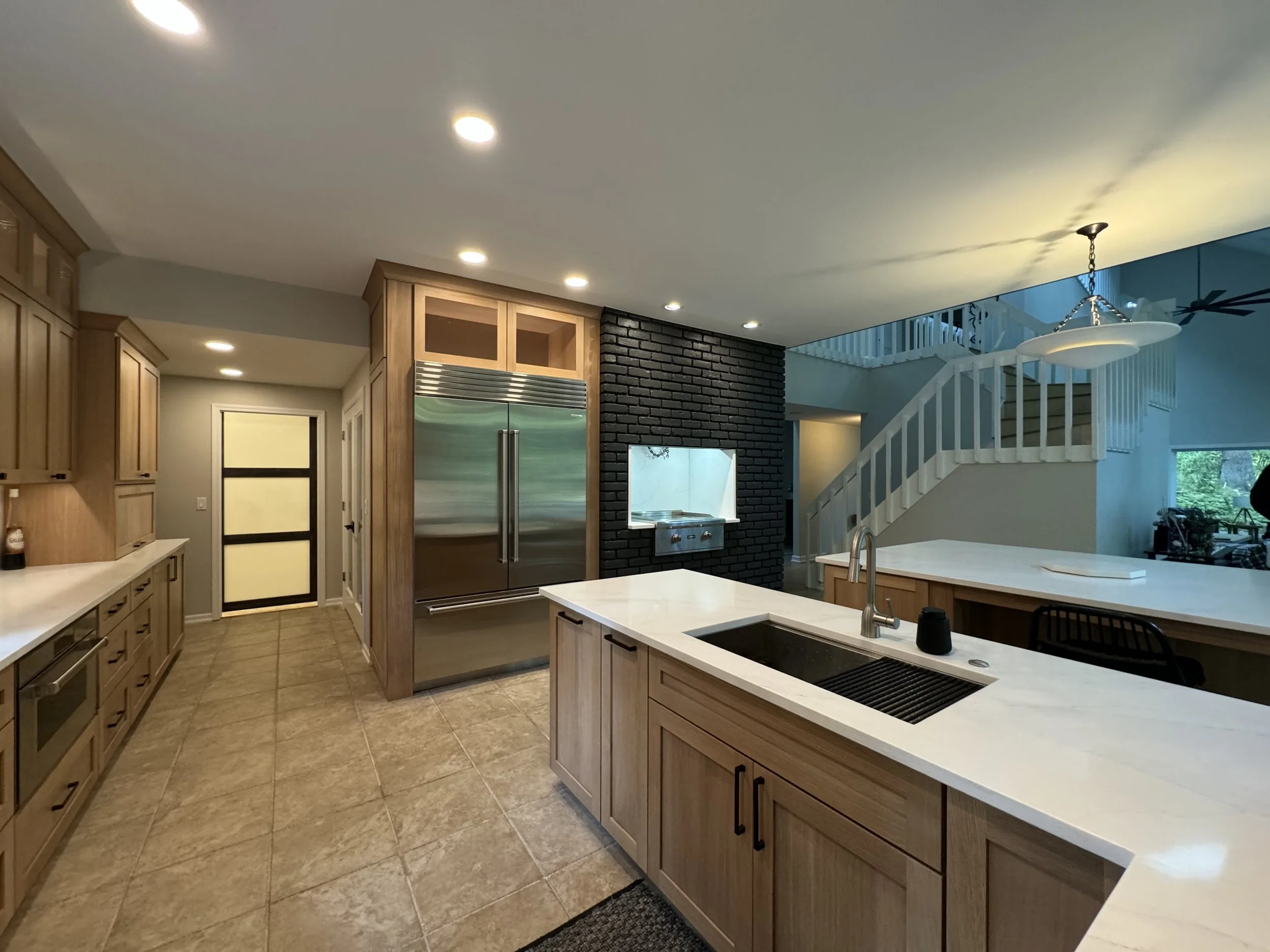 Modern kitchen with wooden cabinets, stainless steel refrigerator, black brick accent wall, white countertop island with sink, tiled floor, and a staircase with white railing in the background.