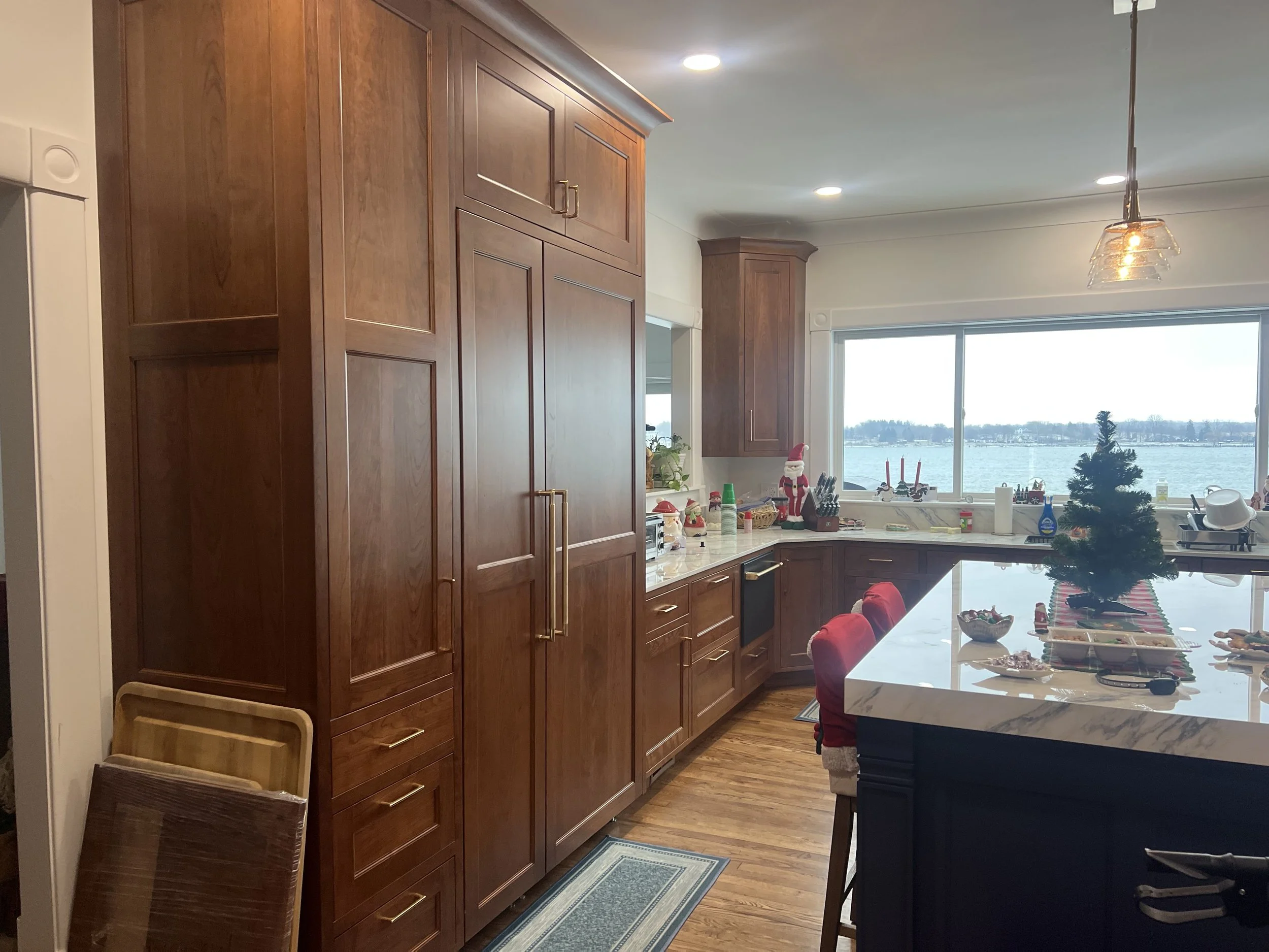A view of a kitchen with wooden cabinets, a white marble island, and holiday decorations including a small Christmas tree, a Santa figurine, and holiday-themed items on the counter. There is a large window showing a water view outside.