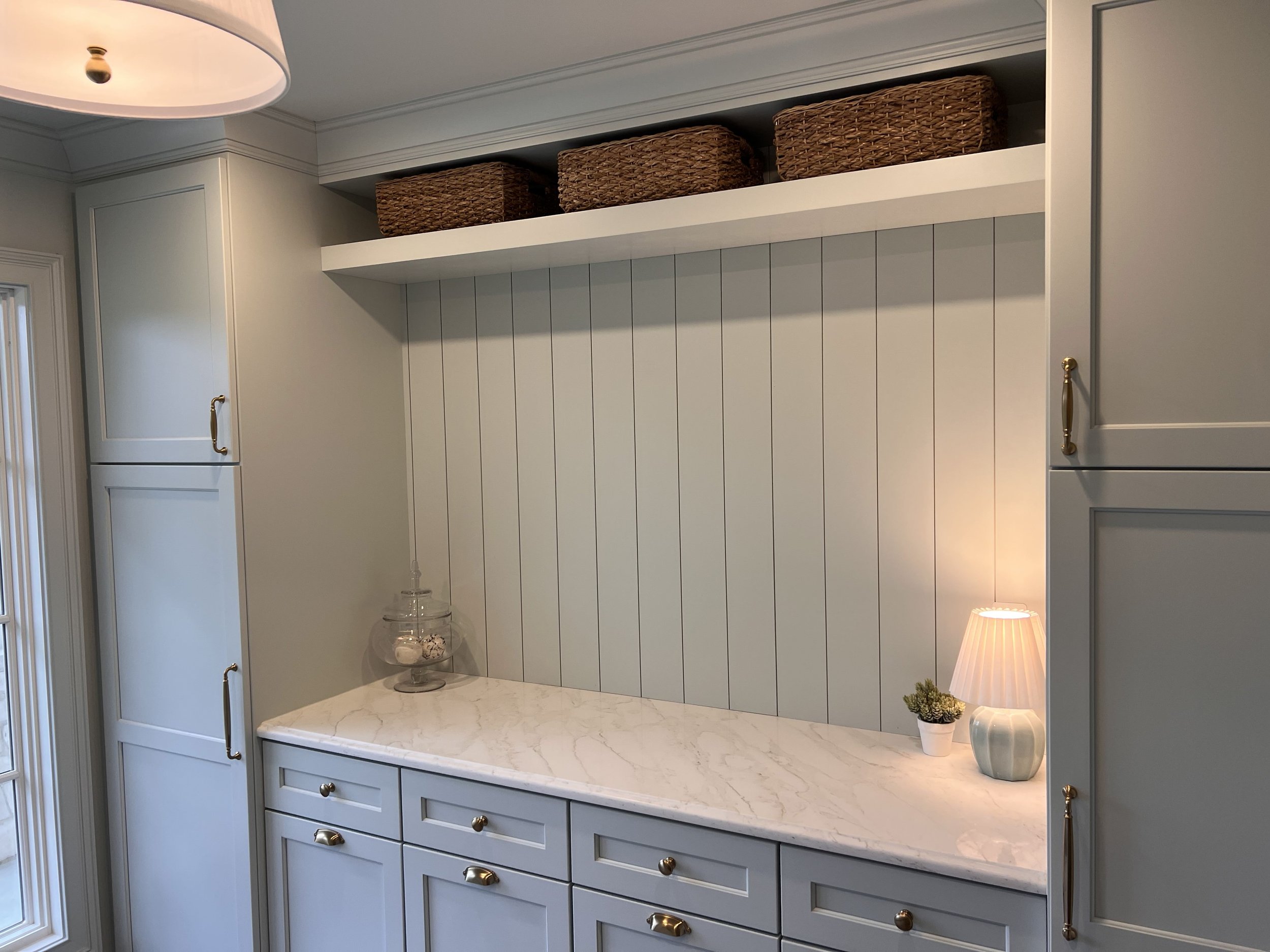 A kitchen countertop with a marble surface, a decorative glass container, a small potted plant, and a lamp with a pleated shade. There are cabinets with gold handles and a backsplash with vertical white panels. Baskets are stored on an upper shelf.