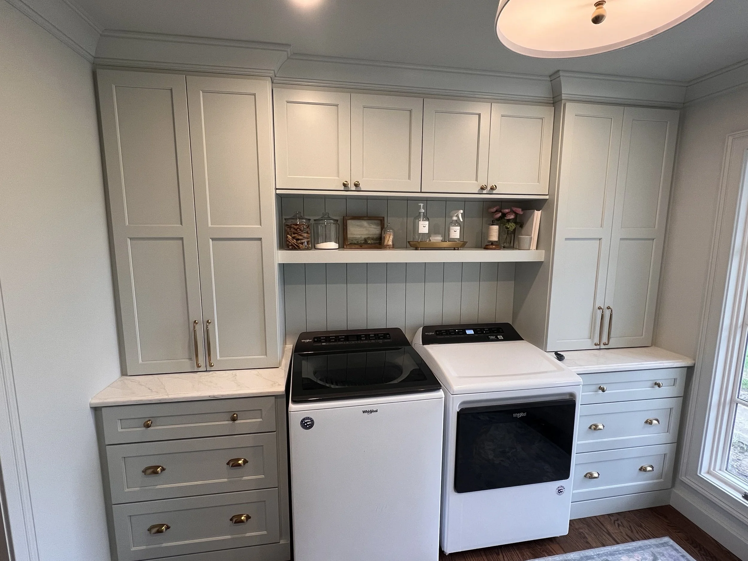 A kitchen with white cabinets, a white marble countertop, and a white black stove. The cabinets have gold knobs, and there are various decorative bottles and boxes on the shelf above the stove. A window is visible on the right side.