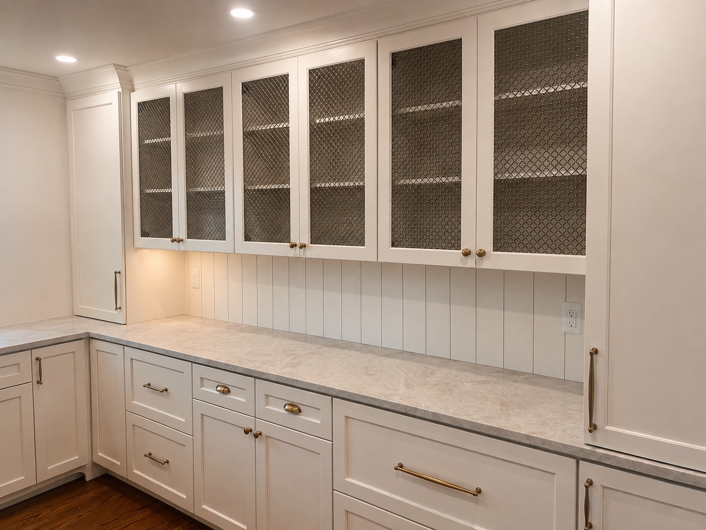White Birmingham MI kitchen cabinets with gold handles, glass doors with black metal mesh panels, beige marble countertop, white beadboard backsplash, under-cabinet lighting, and wood flooring.