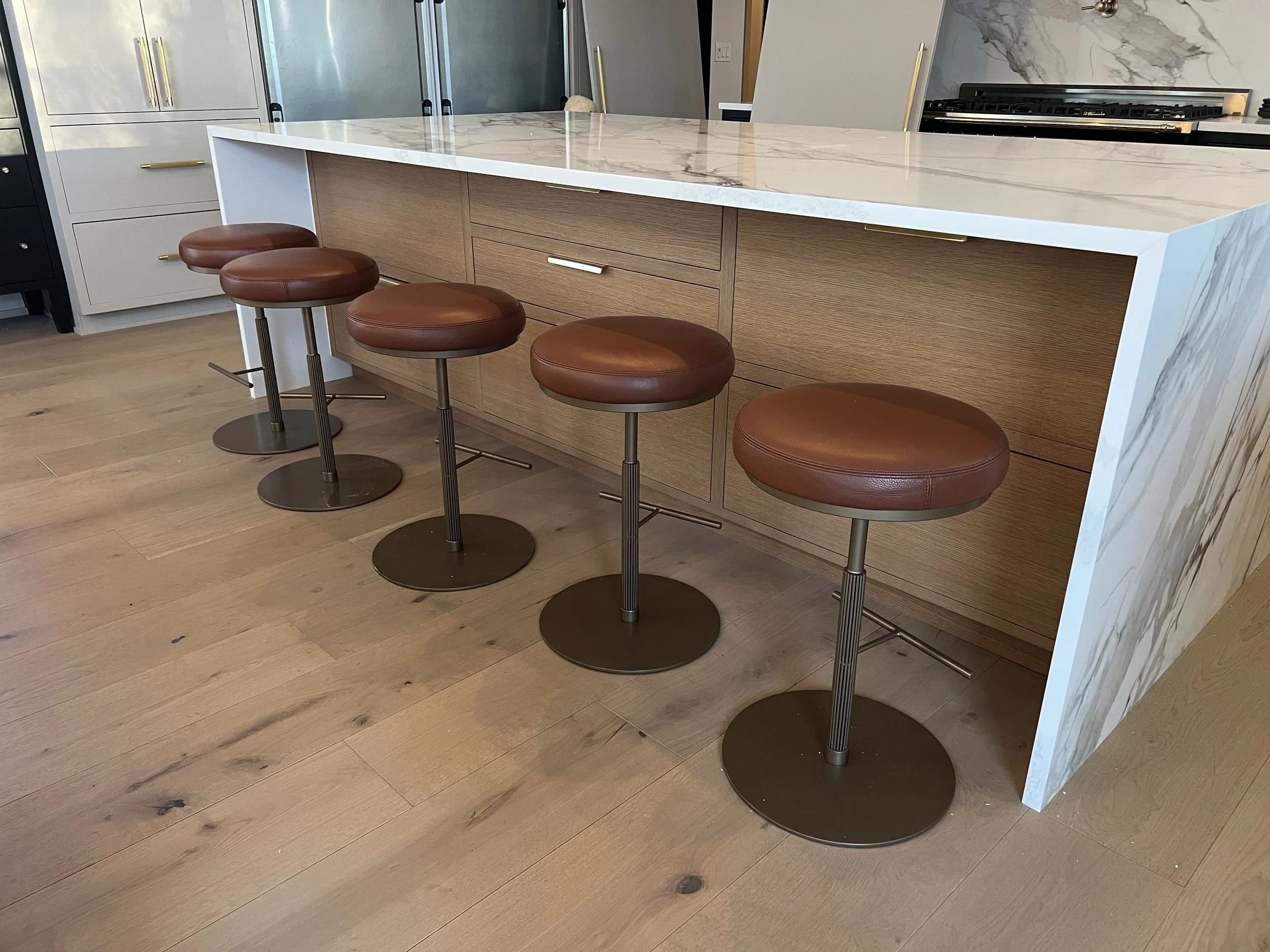 A kitchen with a white marble island, three brown leather bar stools, light wood flooring, and light-colored cabinetry with gold handles.