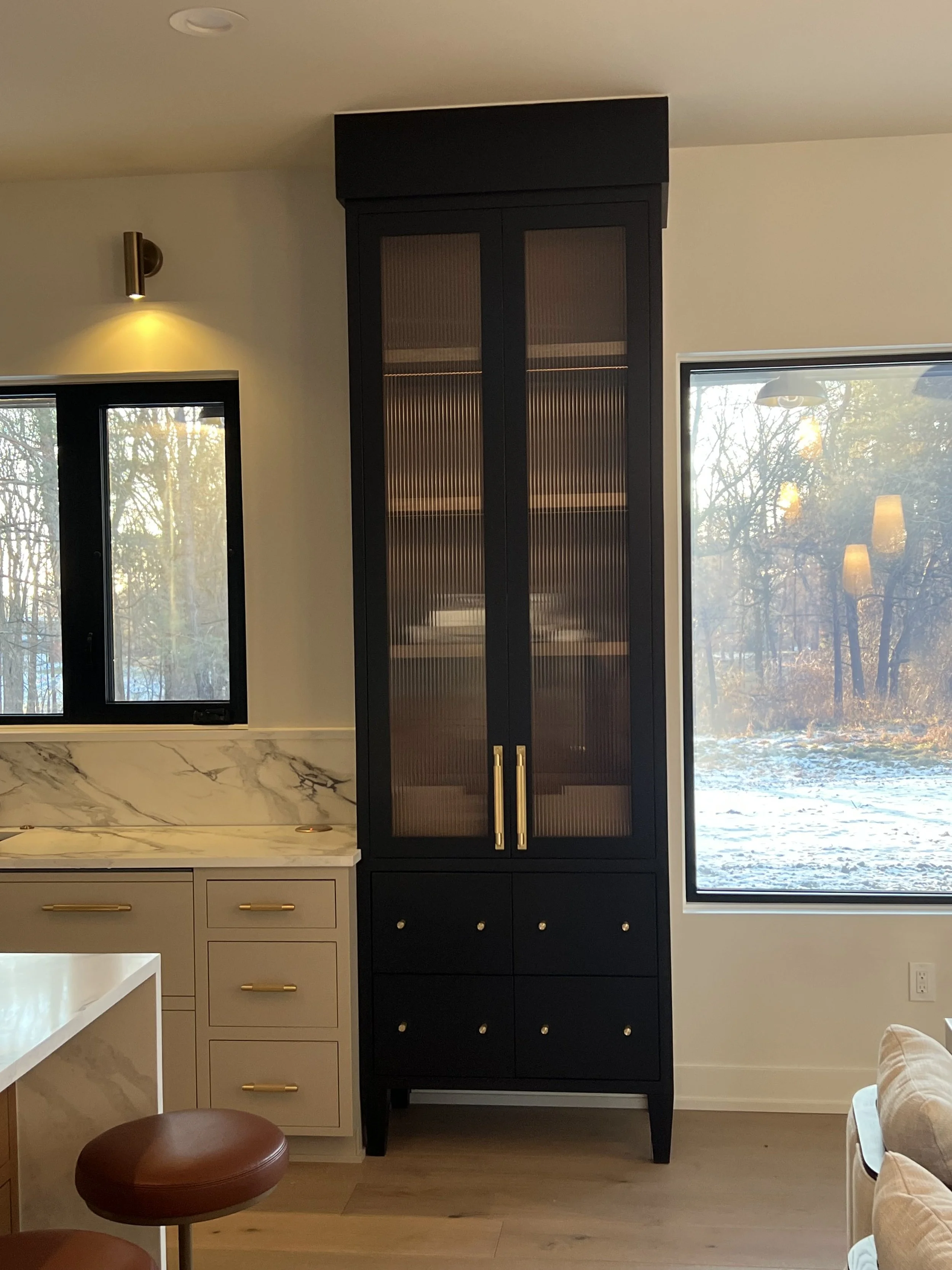Interior view of a kitchen with beige cabinets, marble countertop, black pantry cabinet, and a large window showing a winter landscape outside.